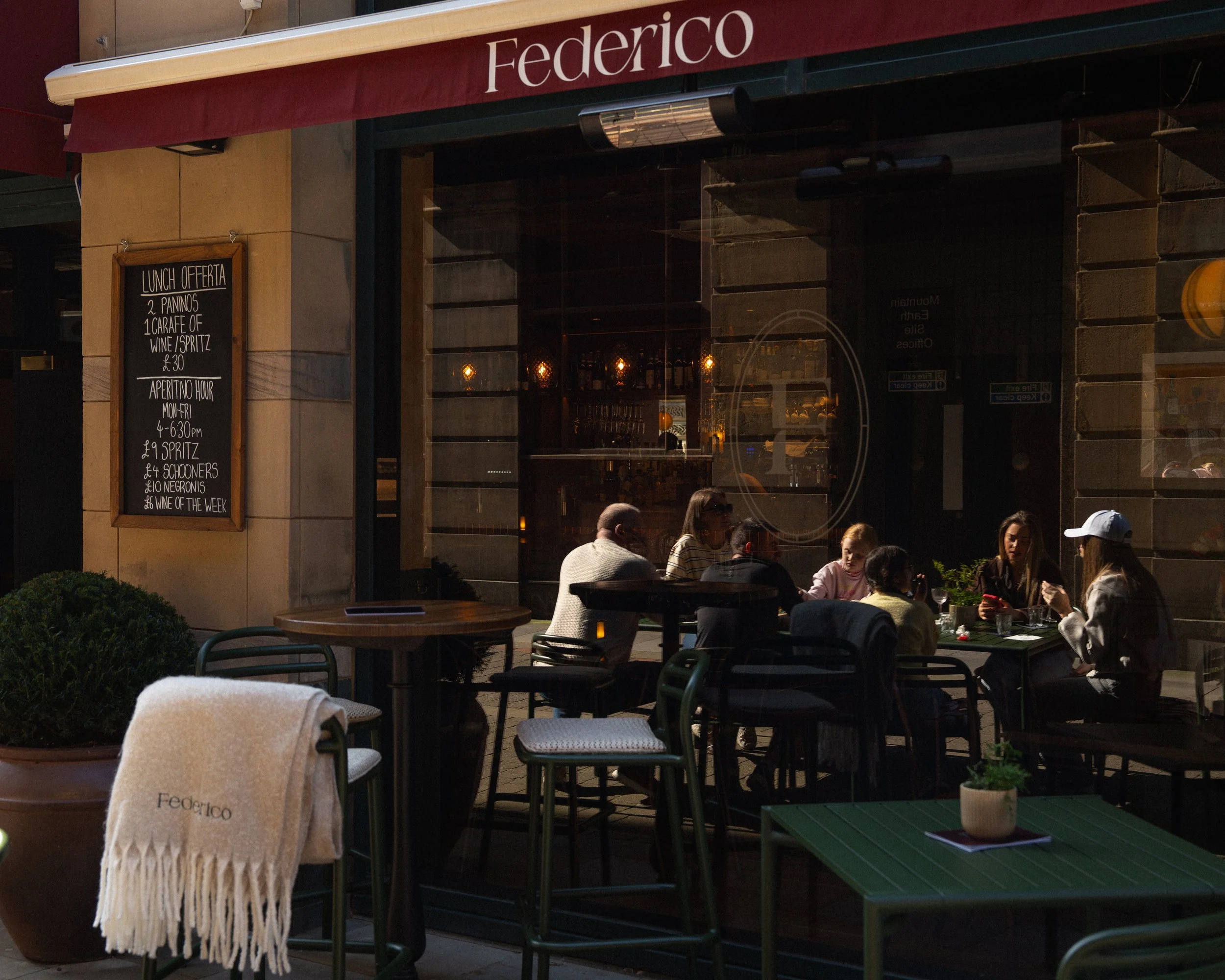 Outside of a restaurant with a red awning labeled 'Federico'. A group of six people are sitting at a table inside, visible through the glass window, enjoying their meal. There are potted plants and a towel with the restaurant name on a chair outside.