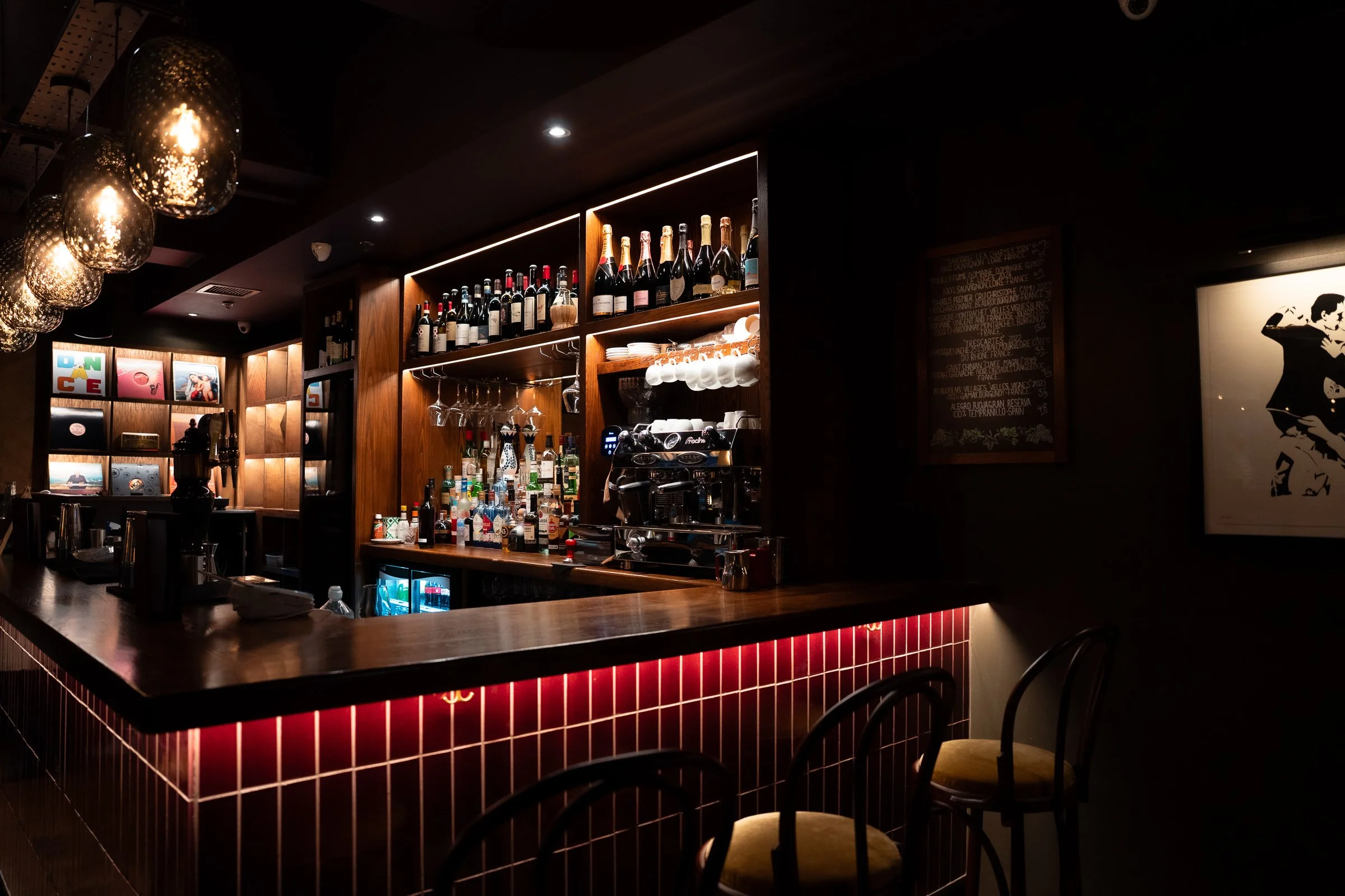 A dimly lit bar with a wooden counter, shelves stocked with various bottles of alcohol and wine, hanging glasses, and a professional espresso machine. Modern light fixtures hang above, and artwork decorates the dark walls.