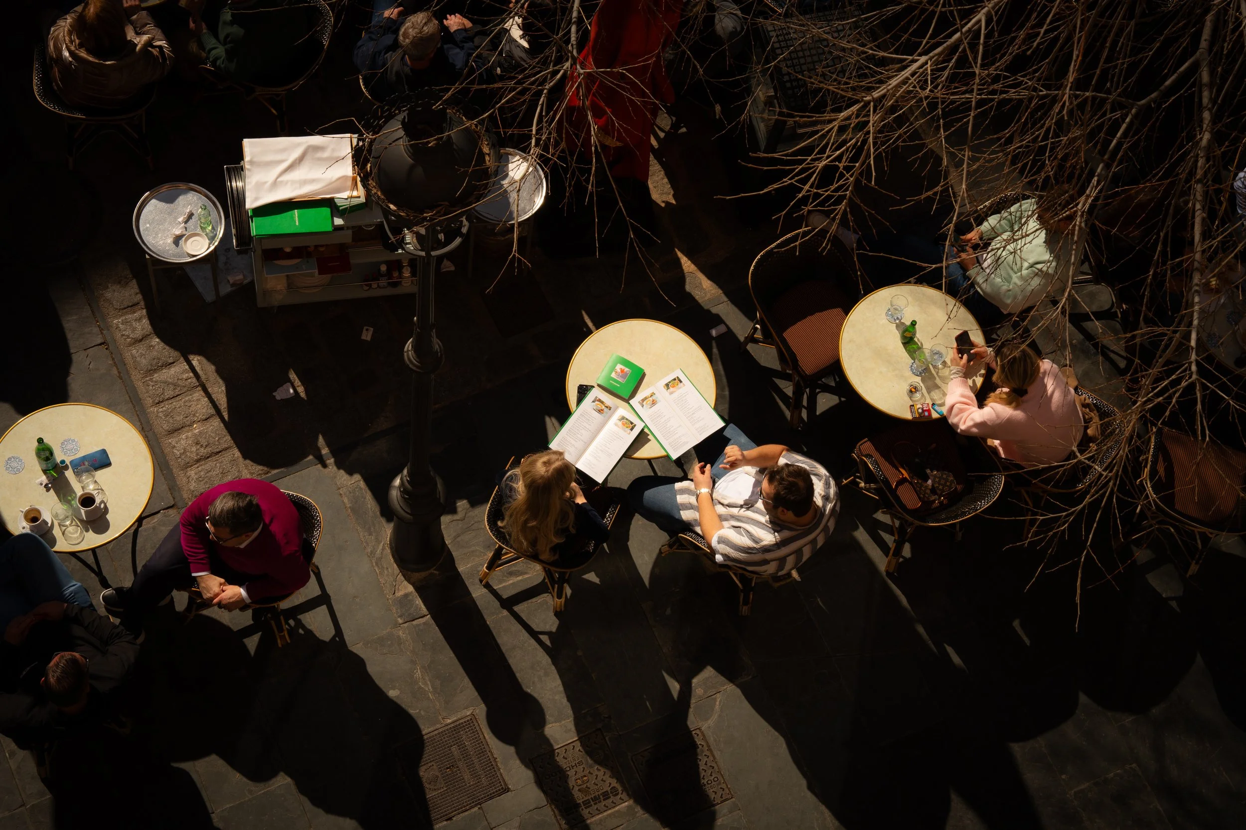 Overhead view of people sitting at outdoor tables at a cafe, with sunlight casting shadows from nearby bare tree branches.
