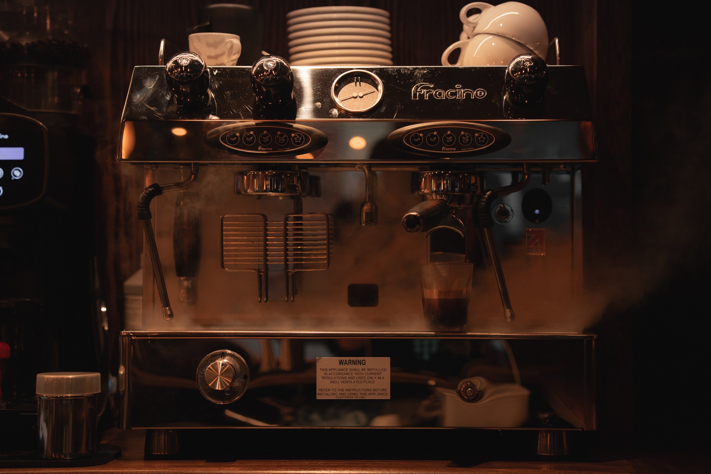A coffee machine with steam rising, topped with cups and saucers, on a dark wooden counter in a coffee shop.