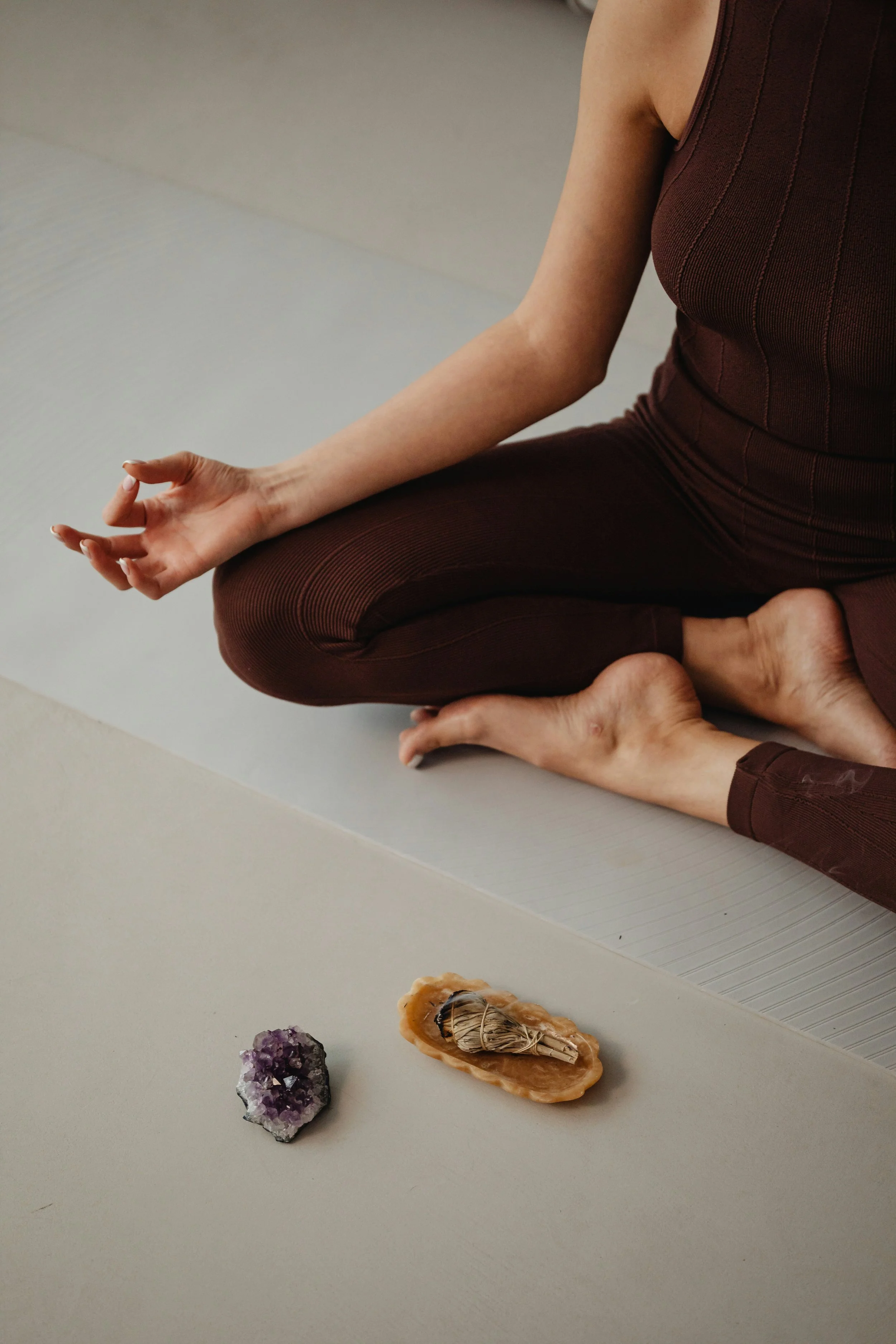 Person meditating in a cross-legged pose on a yoga mat, with two crystals - amethyst and a quartz with a small scroll or bundle - placed in front of them.