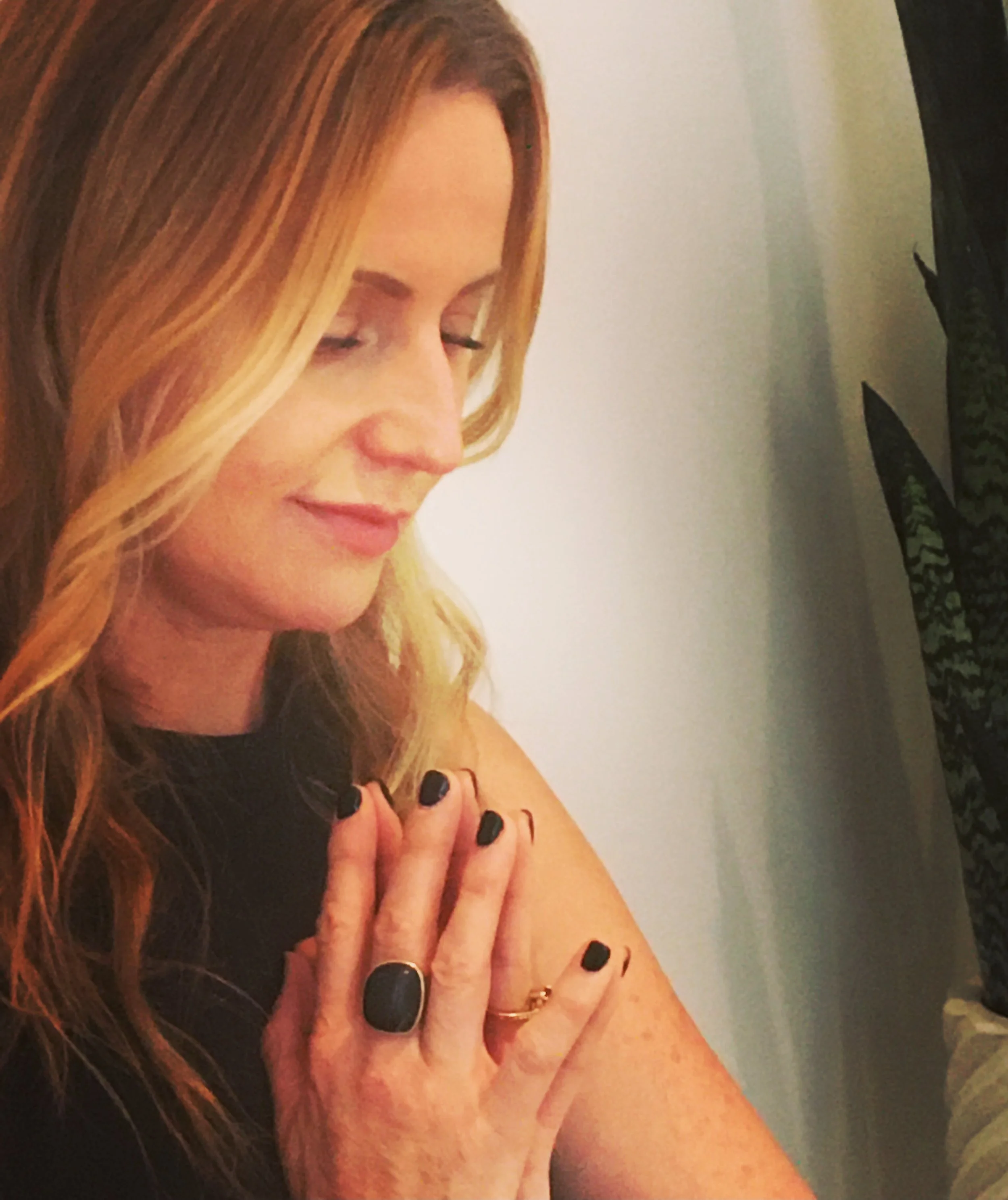 Kiley Efron meditating with closed eyes, and a gentle smile, black nails and a gold ring, against a plain wall with a plant in the background.