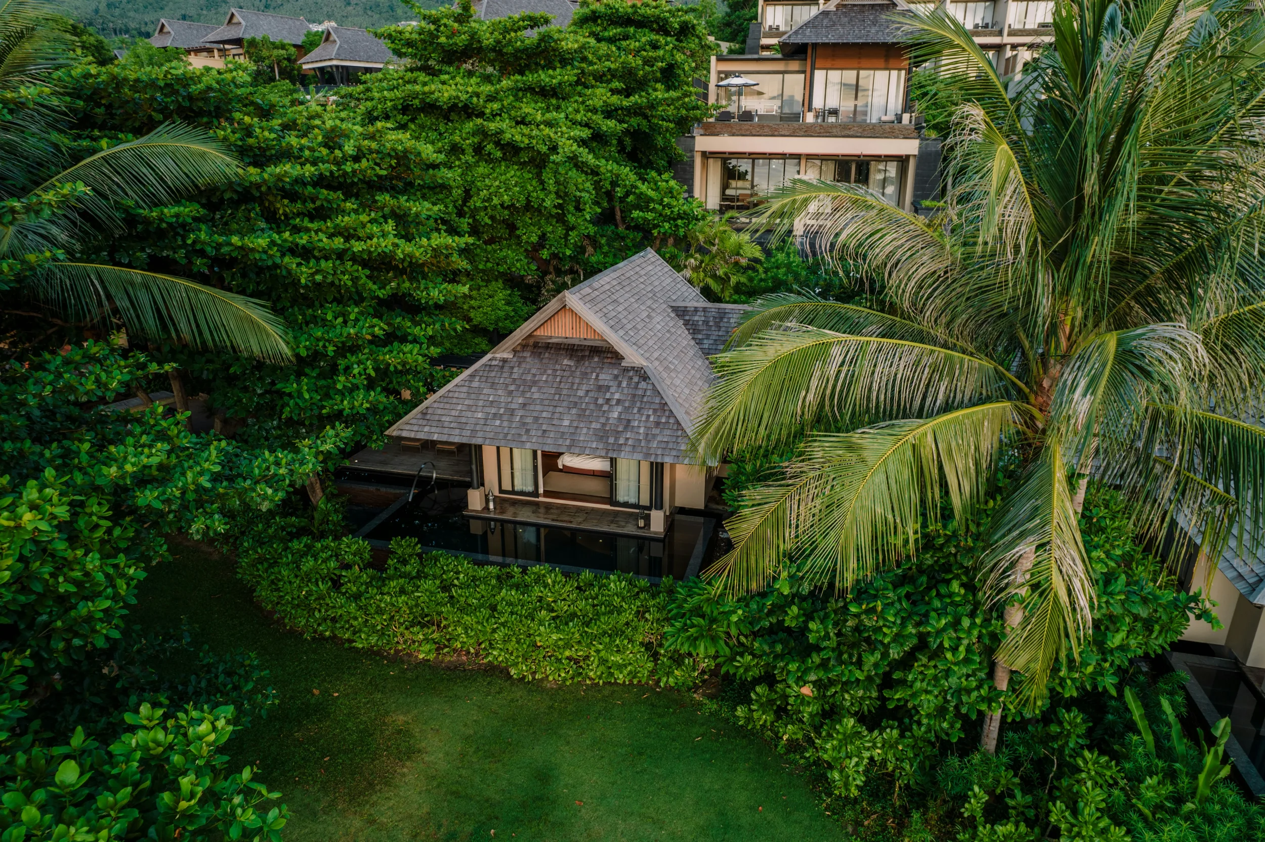 Aerial view of tropical hillside hotel with multiple balconies and lush green trees and palm fronds surrounding it.
