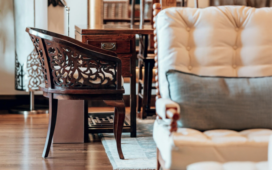 A wooden chair with ornate backrest next to a cream-colored tufted armchair with a pillow, in a room with a wooden floor and a small wooden table.