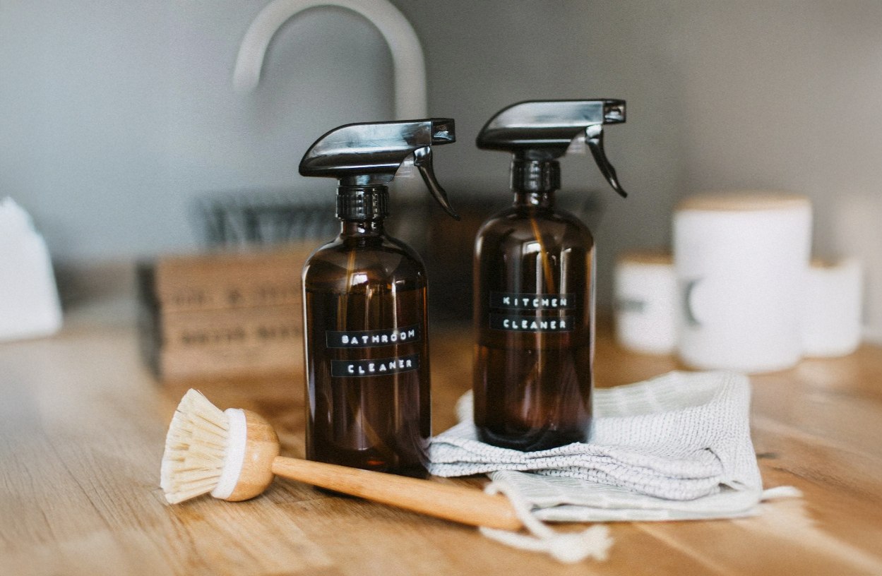 Two brown spray bottles labeled 'Bathroom Cleaner' and 'Kitchen Cleaner' are on a wooden table, accompanied by a cleaning brush and a white towel, with white containers in the background.
