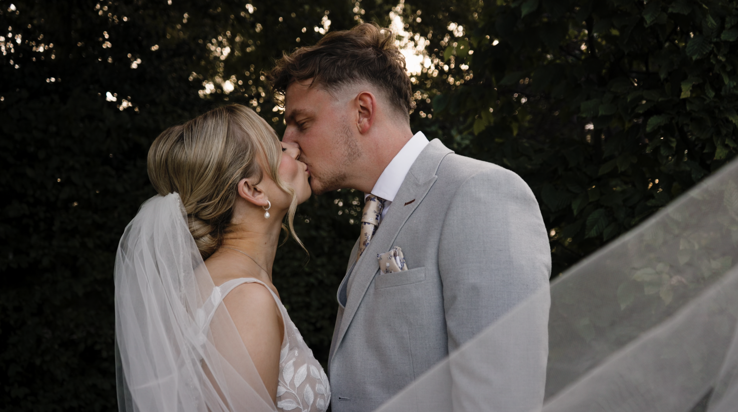 A bride and groom kissing outdoors, surrounded by dark green foliage, during their wedding ceremony.