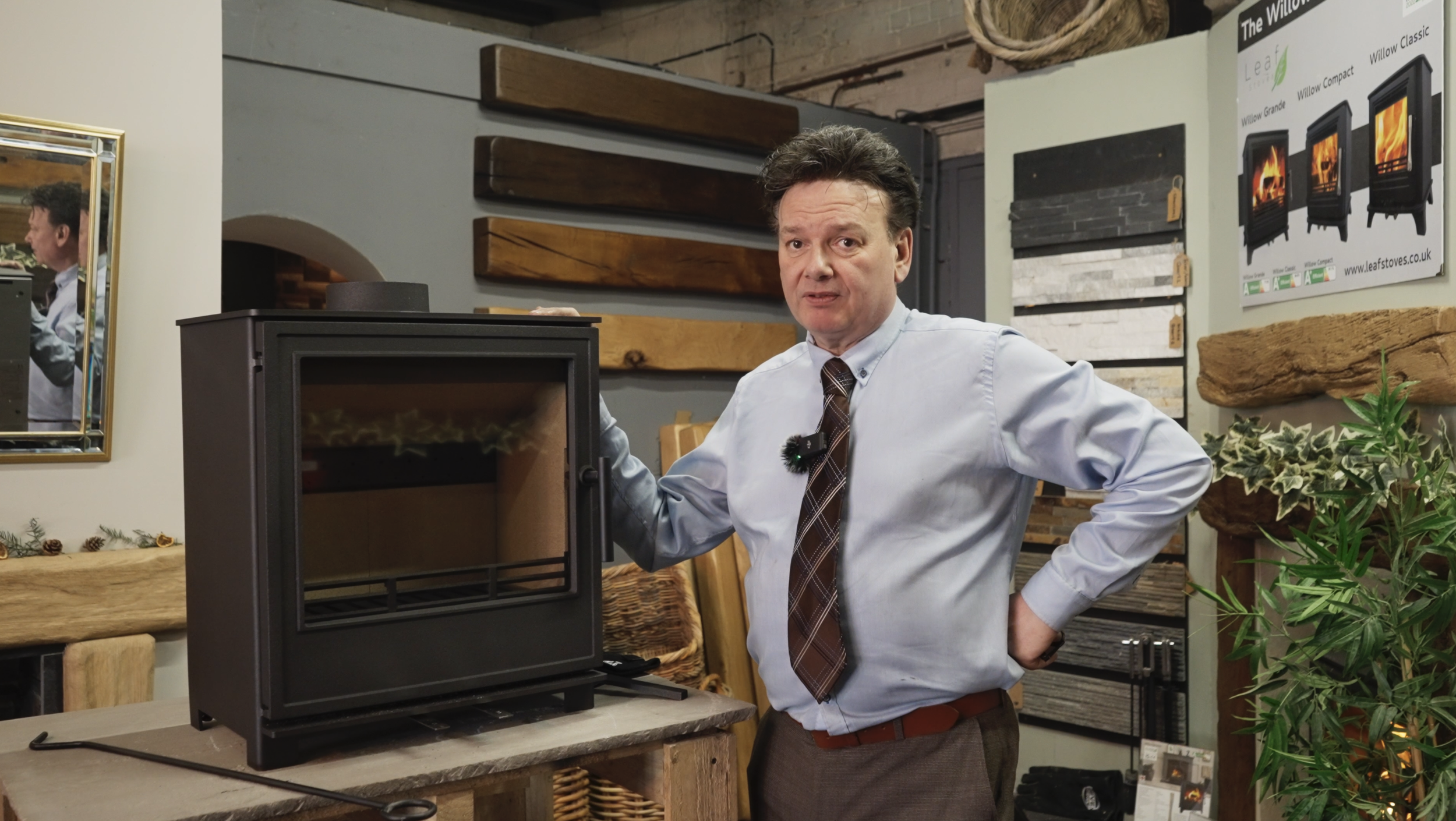 Mark from Hearth & Homes in light blue shirt and plaid tie standing next to a black stove with glass door in a store display area, surrounded by various wooden samples and greenery.
