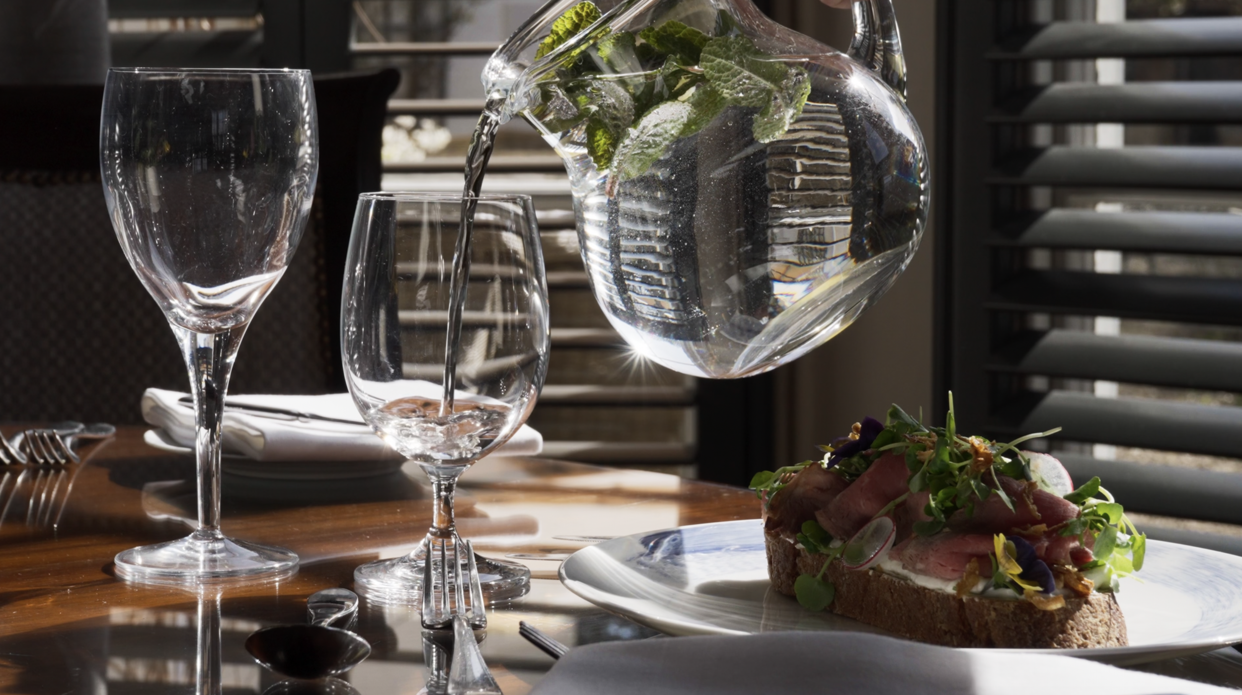 Empty wine glasses, a pitcher of water with mint, and a plate with an open-faced sandwich topped with greens, radishes, and flowers on a wooden table in a sunlit dining setting.