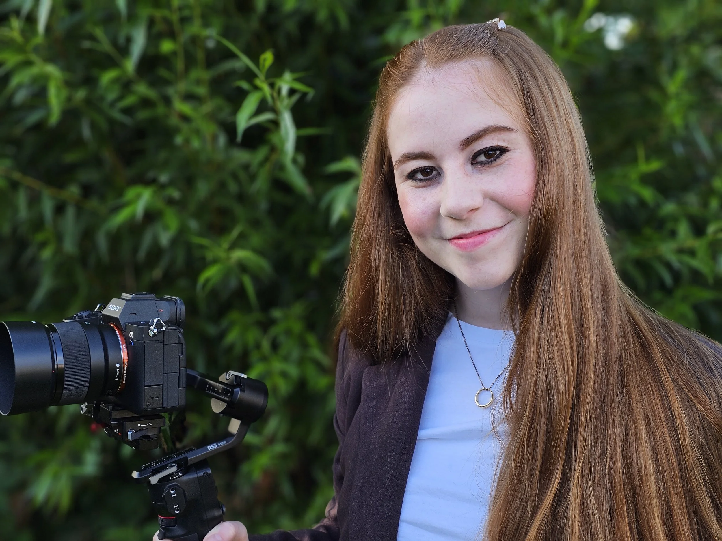 Rachel, the founder of R V Productions with long red hair smiling at camera, standing outdoors near green foliage, holding a professional camera on a gimbal