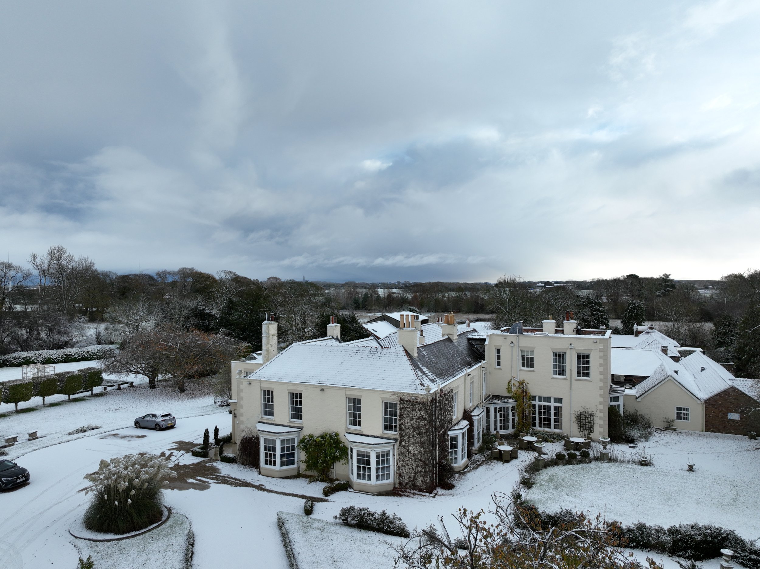 Tickton Grange with snow-covered roof and multiple chimneys, surrounded by a snow-covered yard and trees, under a cloudy sky.