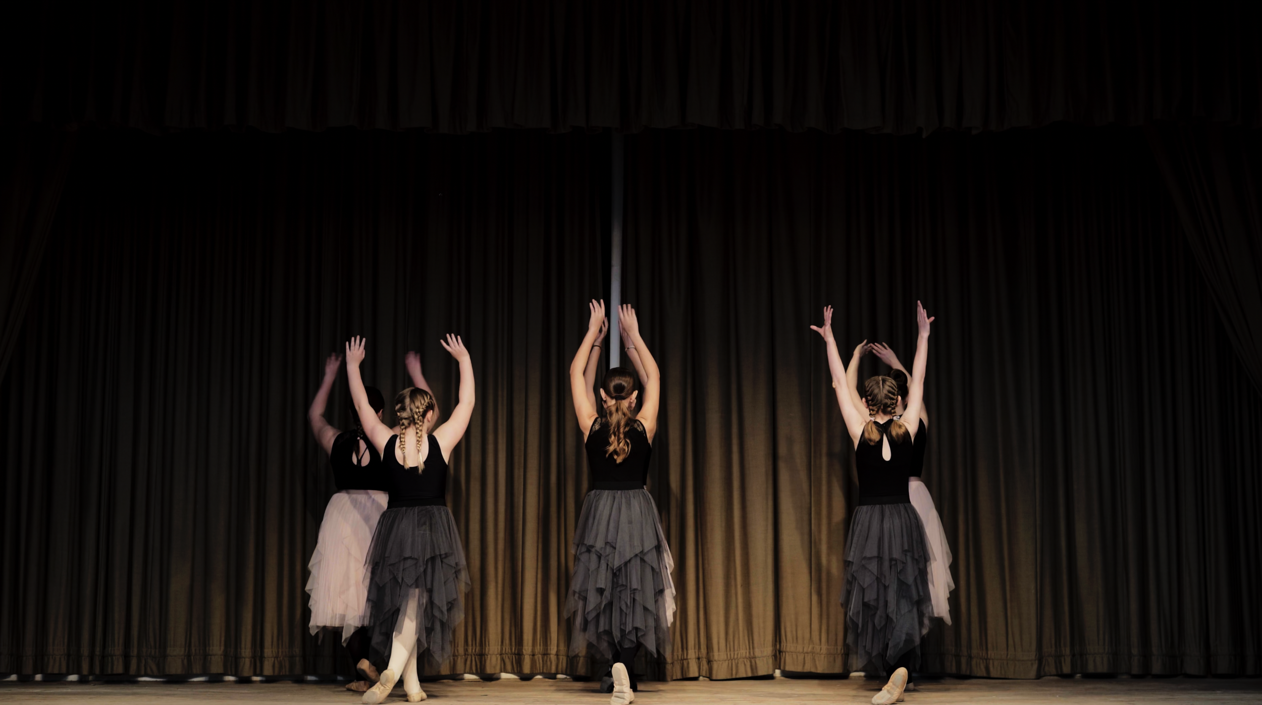 Four ballet dancers on stage perform with arms raised, facing a closed curtain.