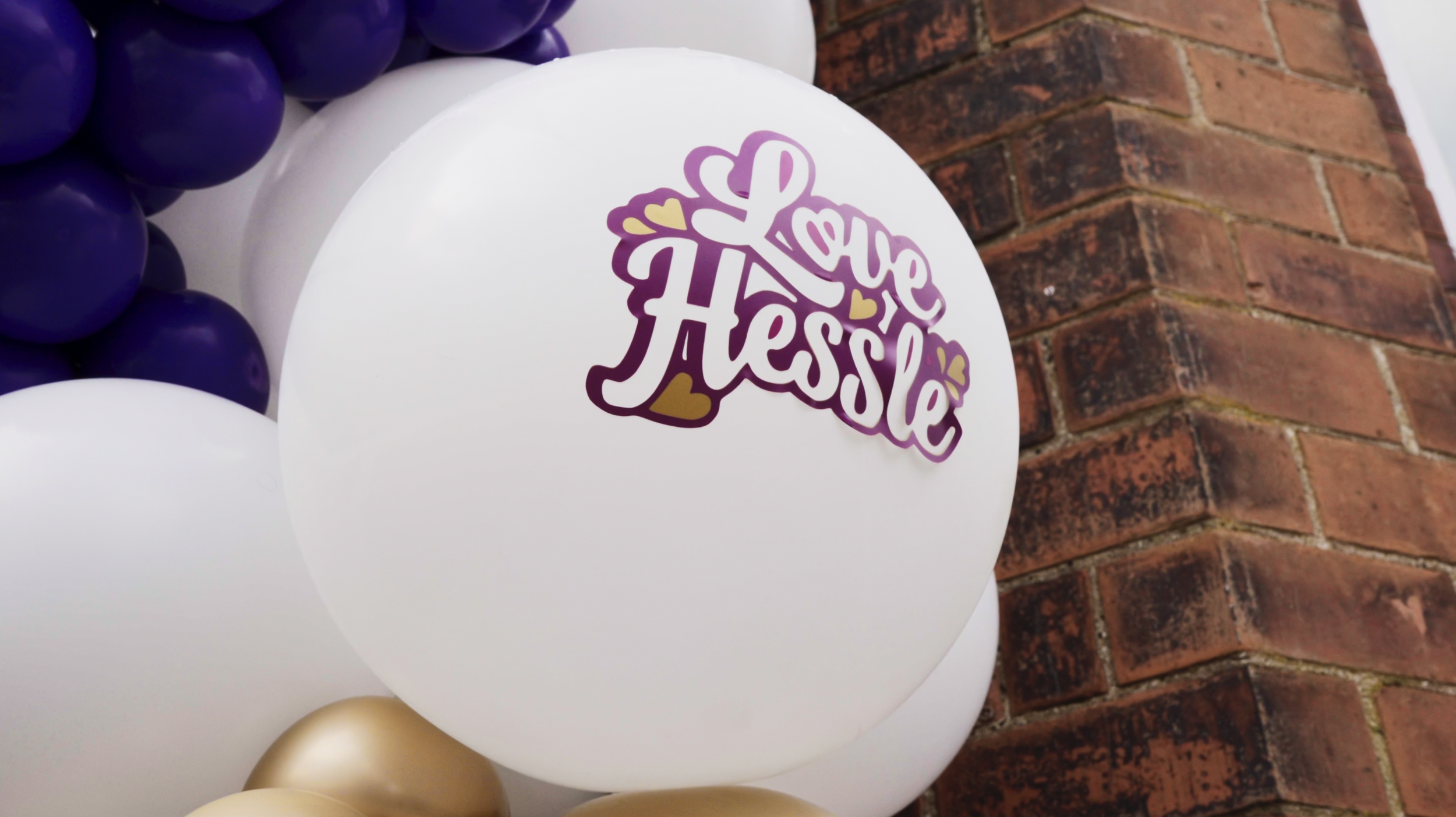 A large white balloon with the words "Love Hessle" written in purple script, surrounded by small yellow hearts, is part of a balloon arrangement next to a brick wall.
