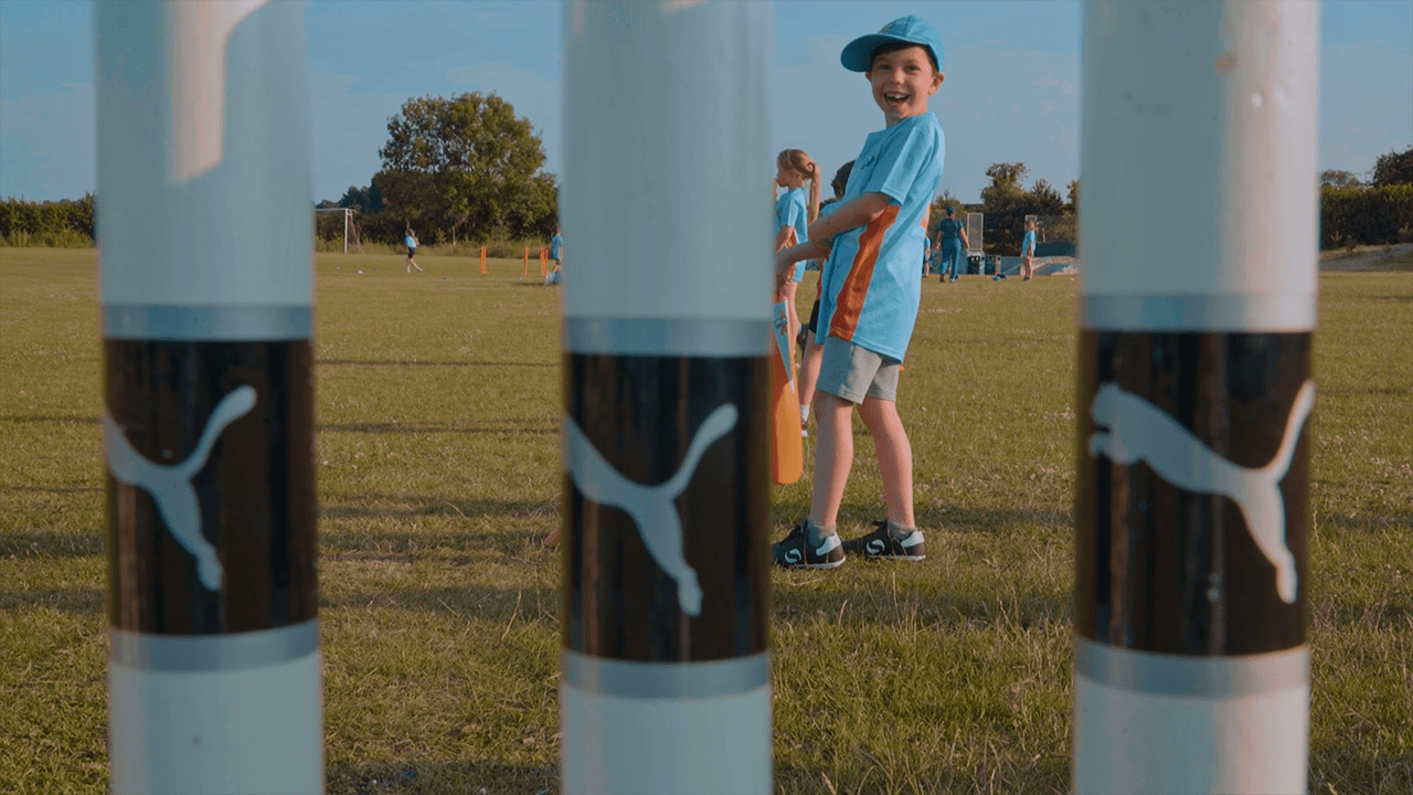 Children playing cricket on a grassy field with soccer goals in the background, viewed through three wickets with Puma logos.
