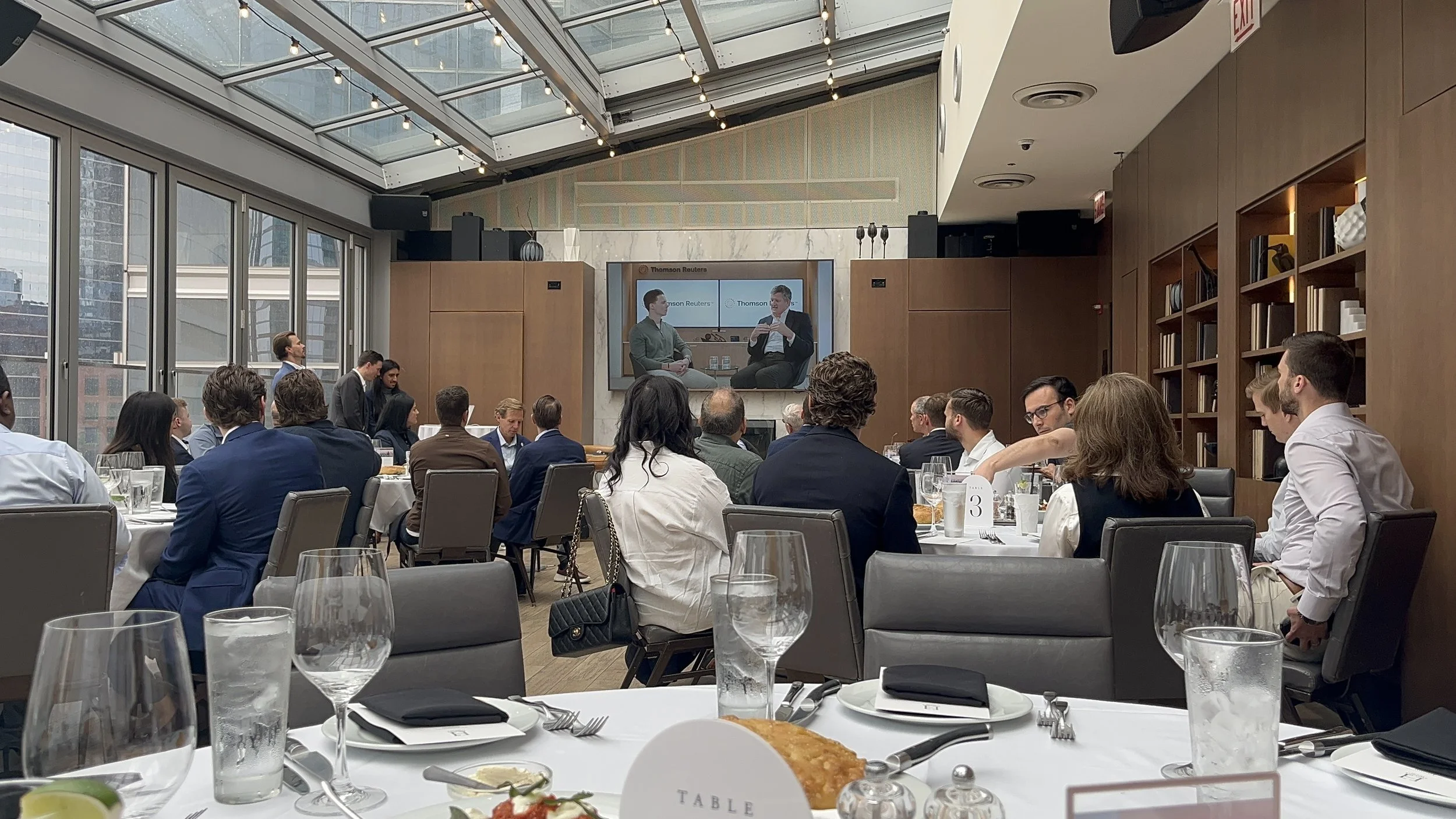 Attendees seated at round tables in a modern conference room with glass ceiling and large windows, listening to a speaker in a TV screen. The room has wood-paneled walls and shelves with books and décor.