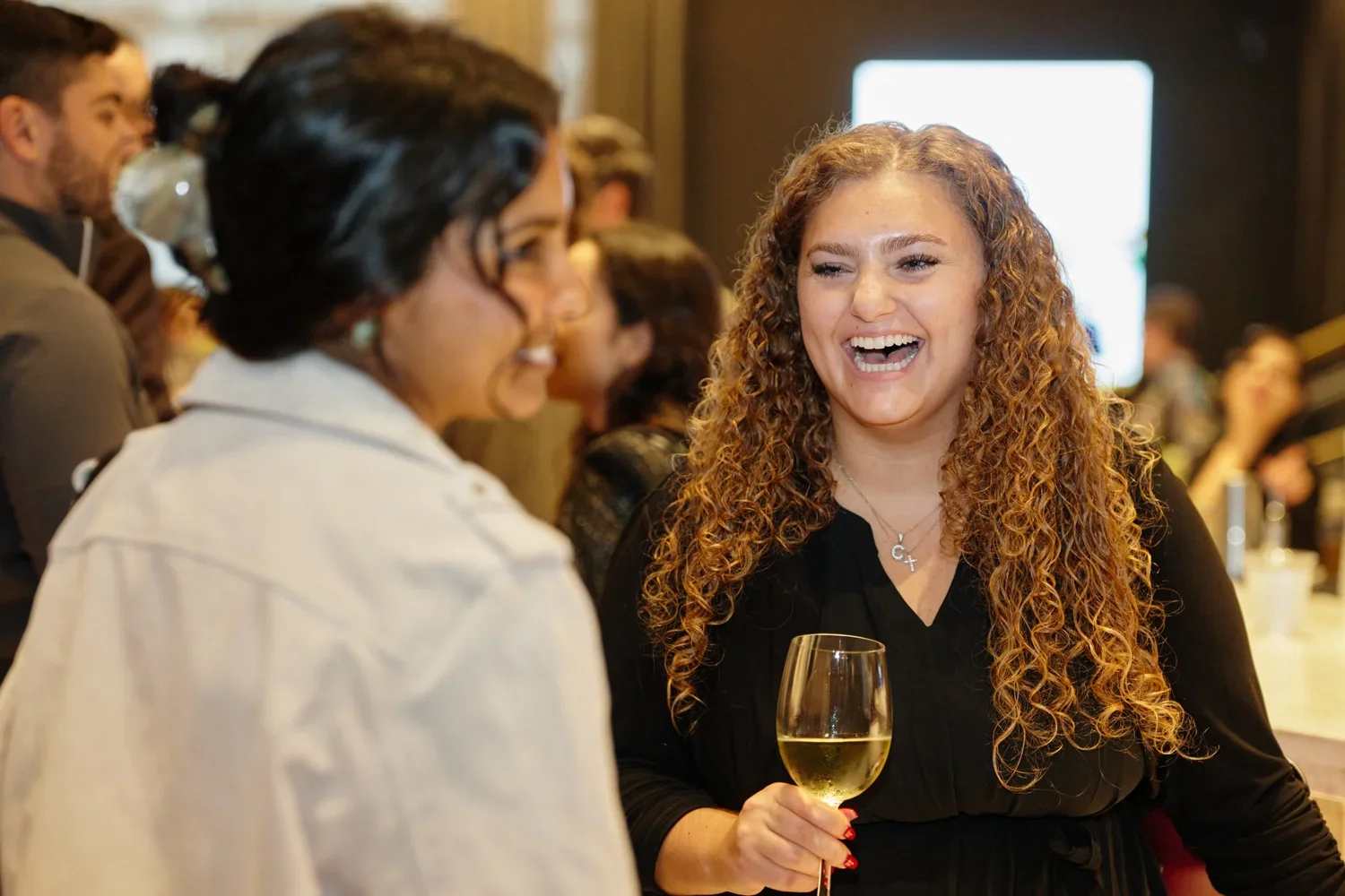 Two women are laughing and talking at a social gathering. The woman on the right has curly red hair, is holding a glass of white wine, and is wearing a black top. The woman on the left has dark hair, glasses, and is wearing a light-colored shirt. Oth