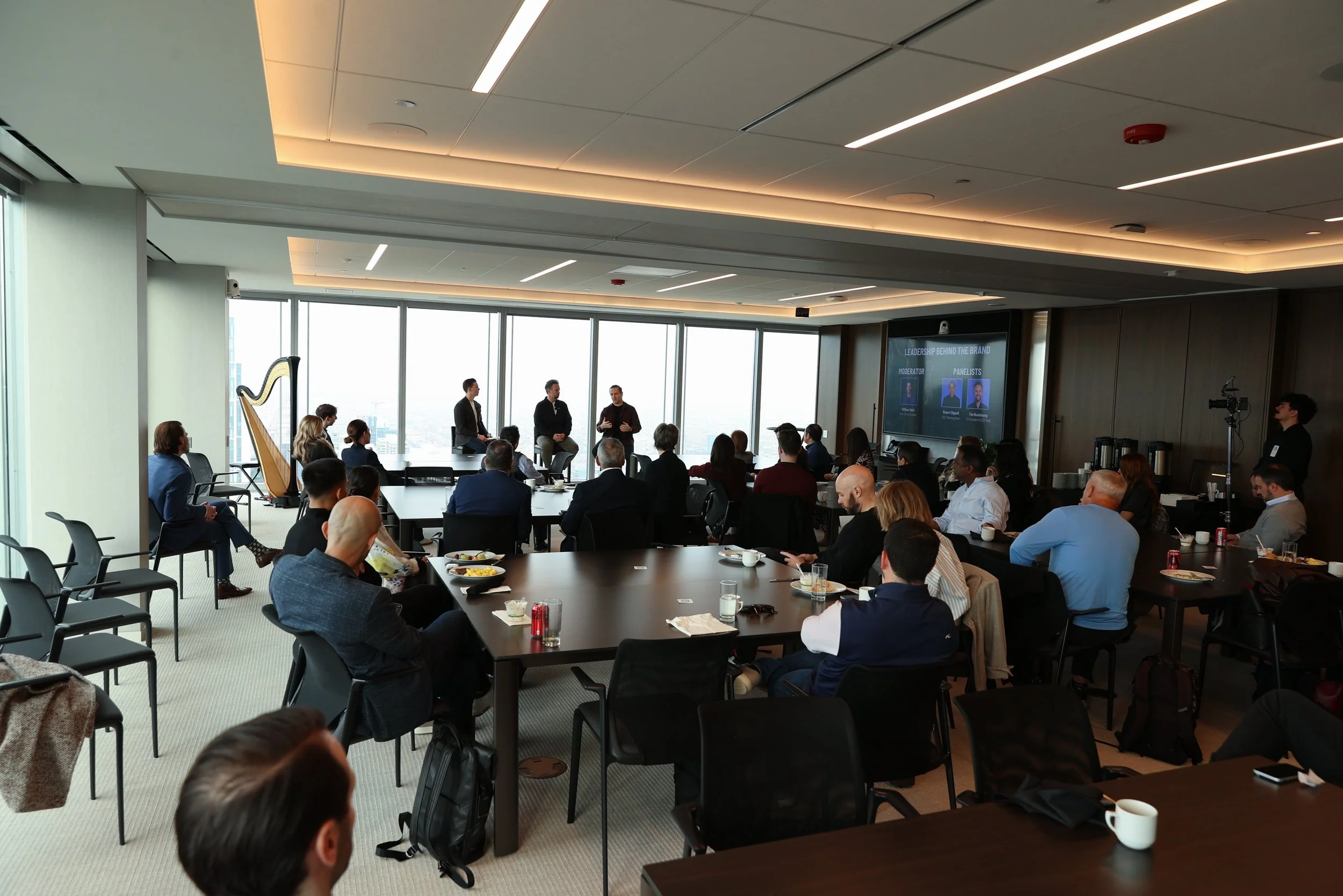 A professional conference with a panel of four speakers on stage in front of large windows overlooking a cityscape. The audience, sitting at tables with refreshments, listens attentively.