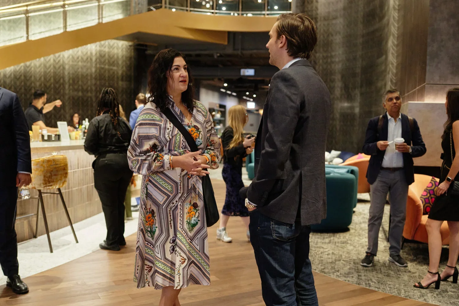 A woman and a man are engaged in conversation in a lobby or lounge area with other people in the background.