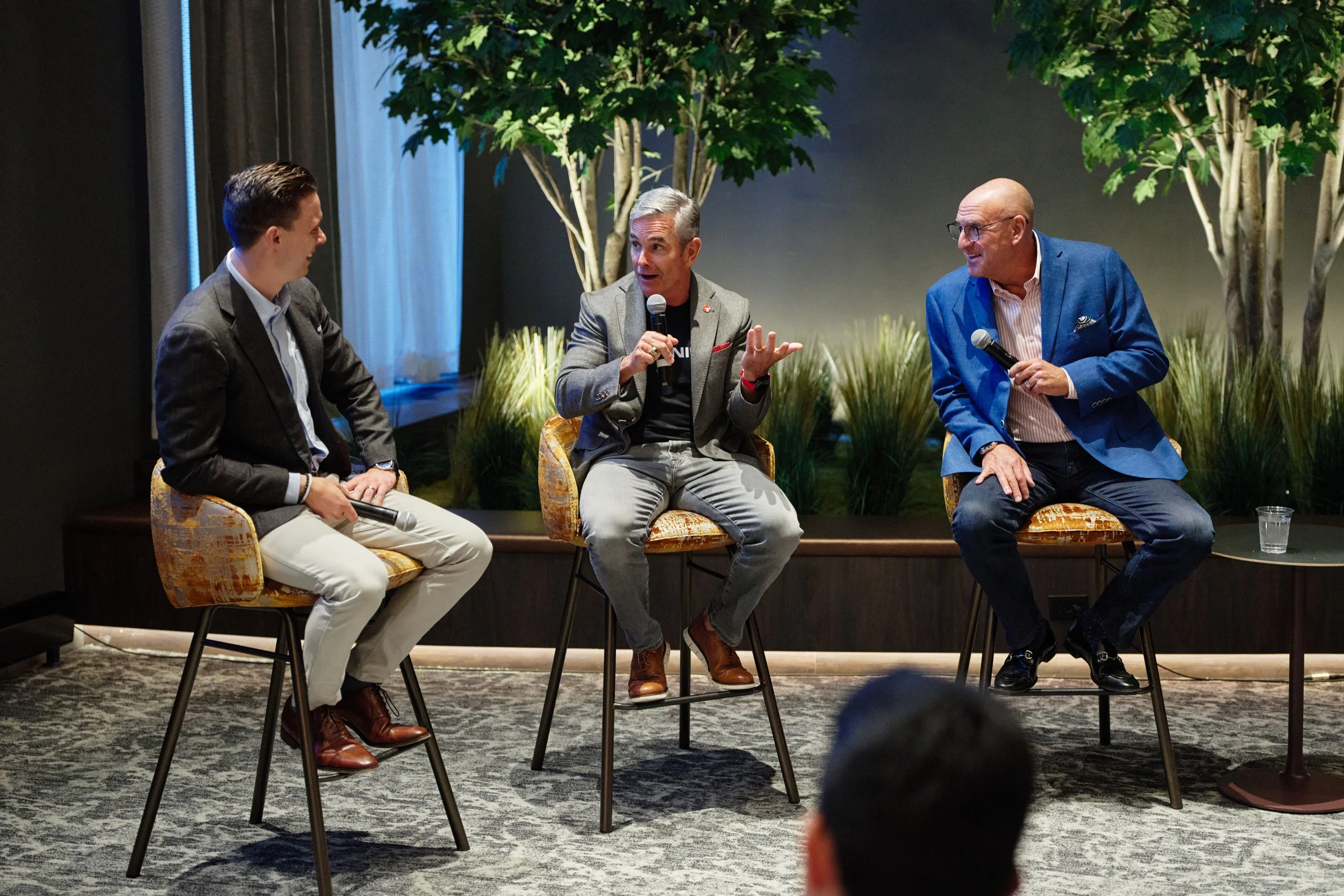 Three men sitting on chairs and engaging in conversation during a panel discussion or interview. They are smiling and holding microphones. The setting has a large potted plant in the background and a dark wall.