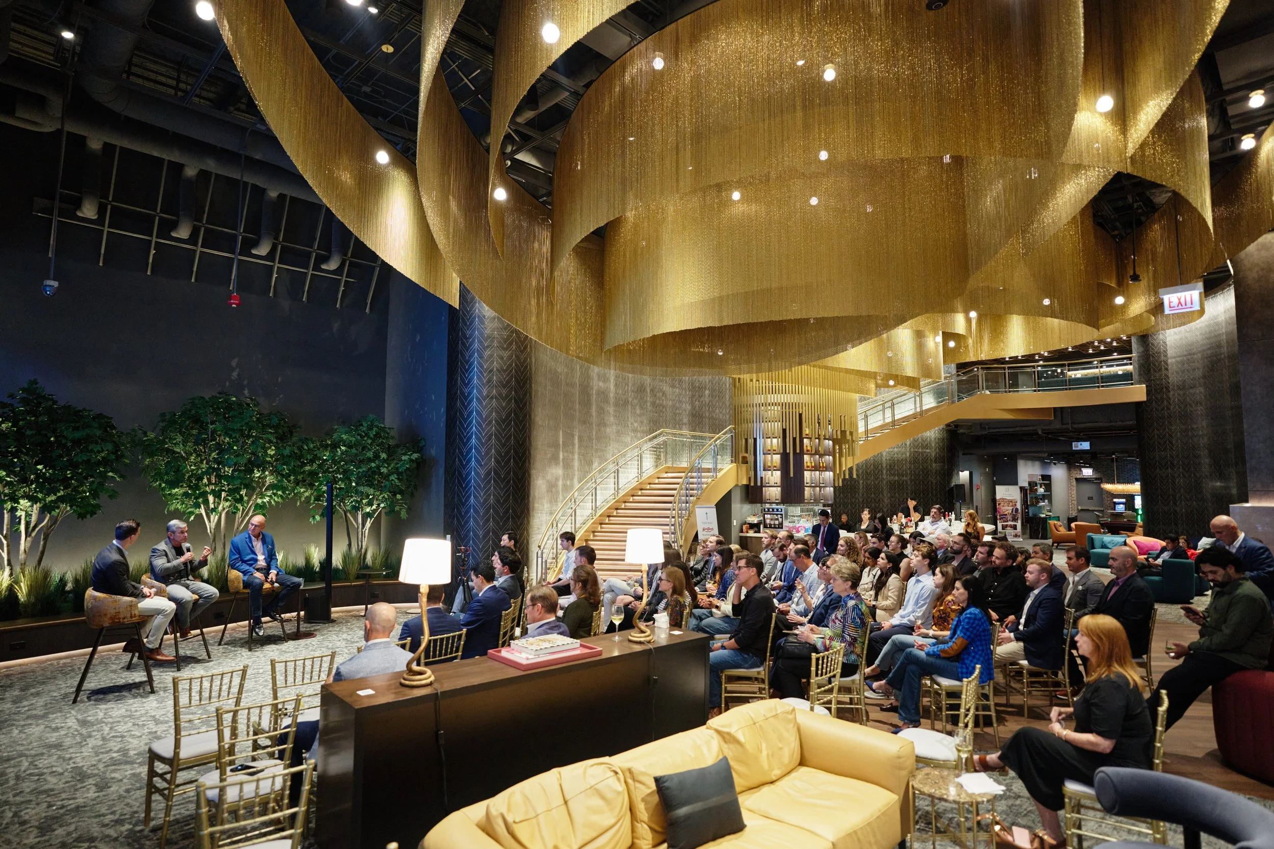 A large crowd gathering in an upscale indoor venue with a gold swirling ceiling decoration, a staircase, and various seating areas. Several people are seated and some are engaged in discussion, indicating an event or conference.