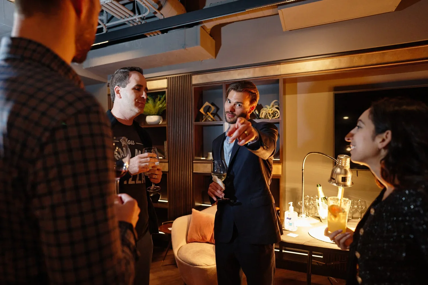 Four people socializing in a modern indoor setting with drinks in hand, one man in a suit pointing towards the camera.