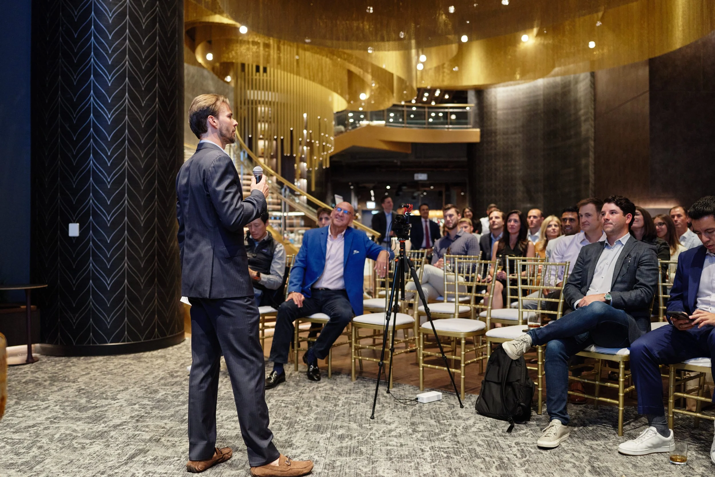 A man in a pinstripe suit holding a microphone and speaking to an audience seated in a formal setting with modern gold and black decor.