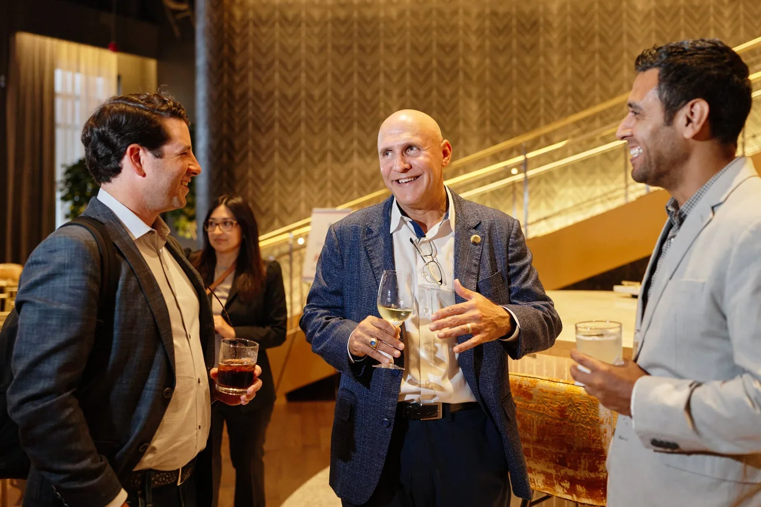Three men and one woman at a social event, with the men holding drinks, engaged in conversation and smiling, in a warm, stylish indoor setting.