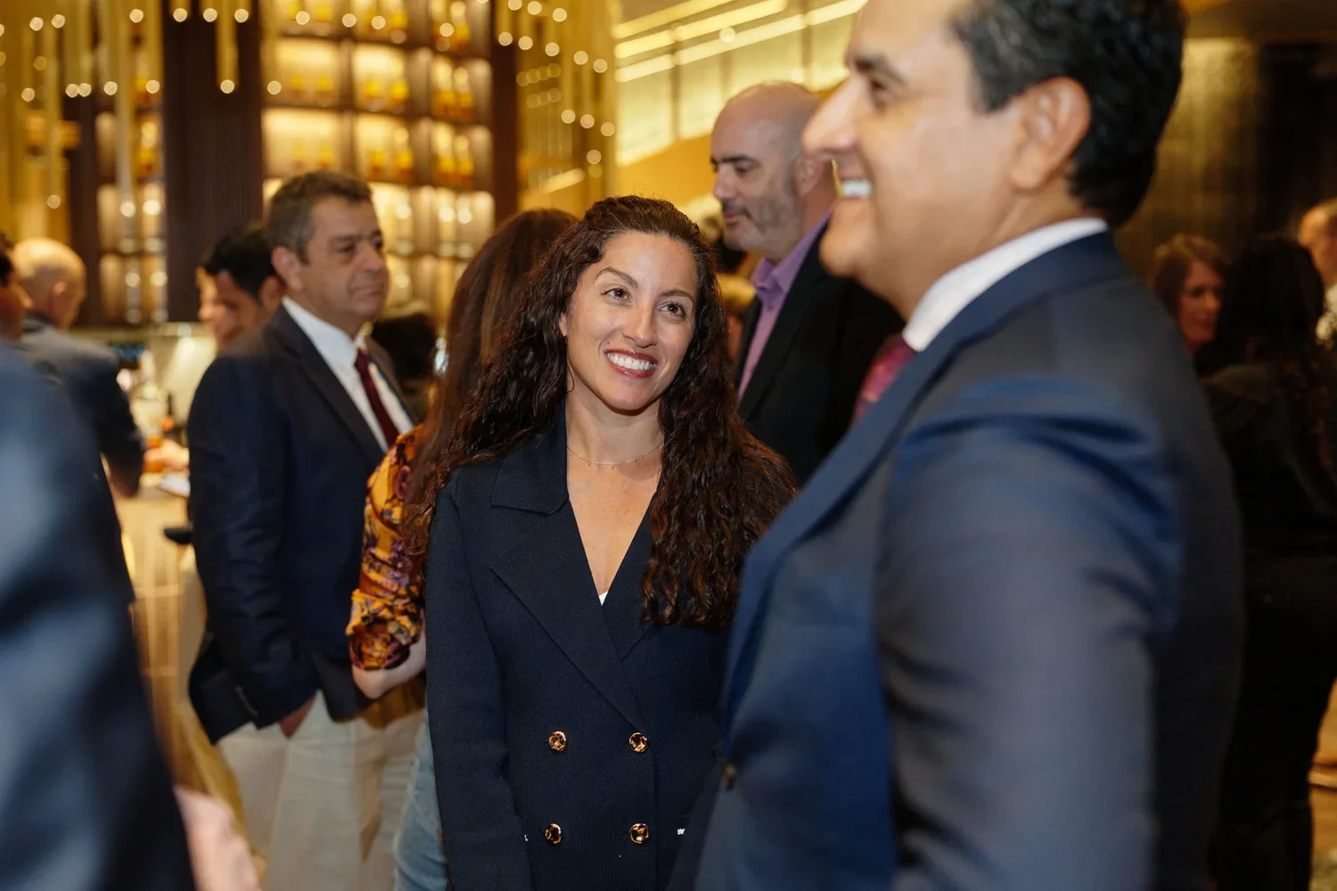 People networking at a professional event or conference, with a woman smiling and engaging in conversation with a man in a suit, surrounded by other attendees in a modern, well-lit venue.