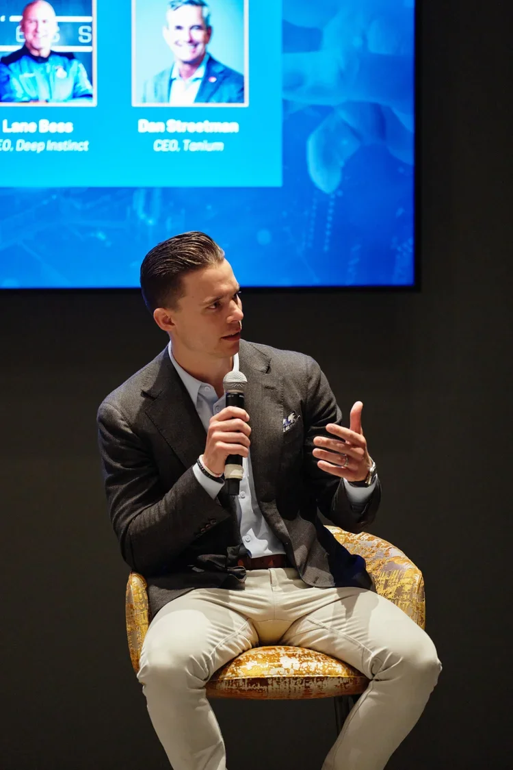 A man in a gray blazer and beige pants holding a microphone, sitting on a yellow patterned chair, speaking at a panel discussion or conference.