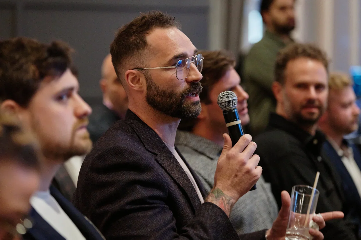 A man in glasses and a dark jacket holding a microphone and a glass of drink, participating in a discussion or Q&A session at a conference or event, surrounded by other attendees.