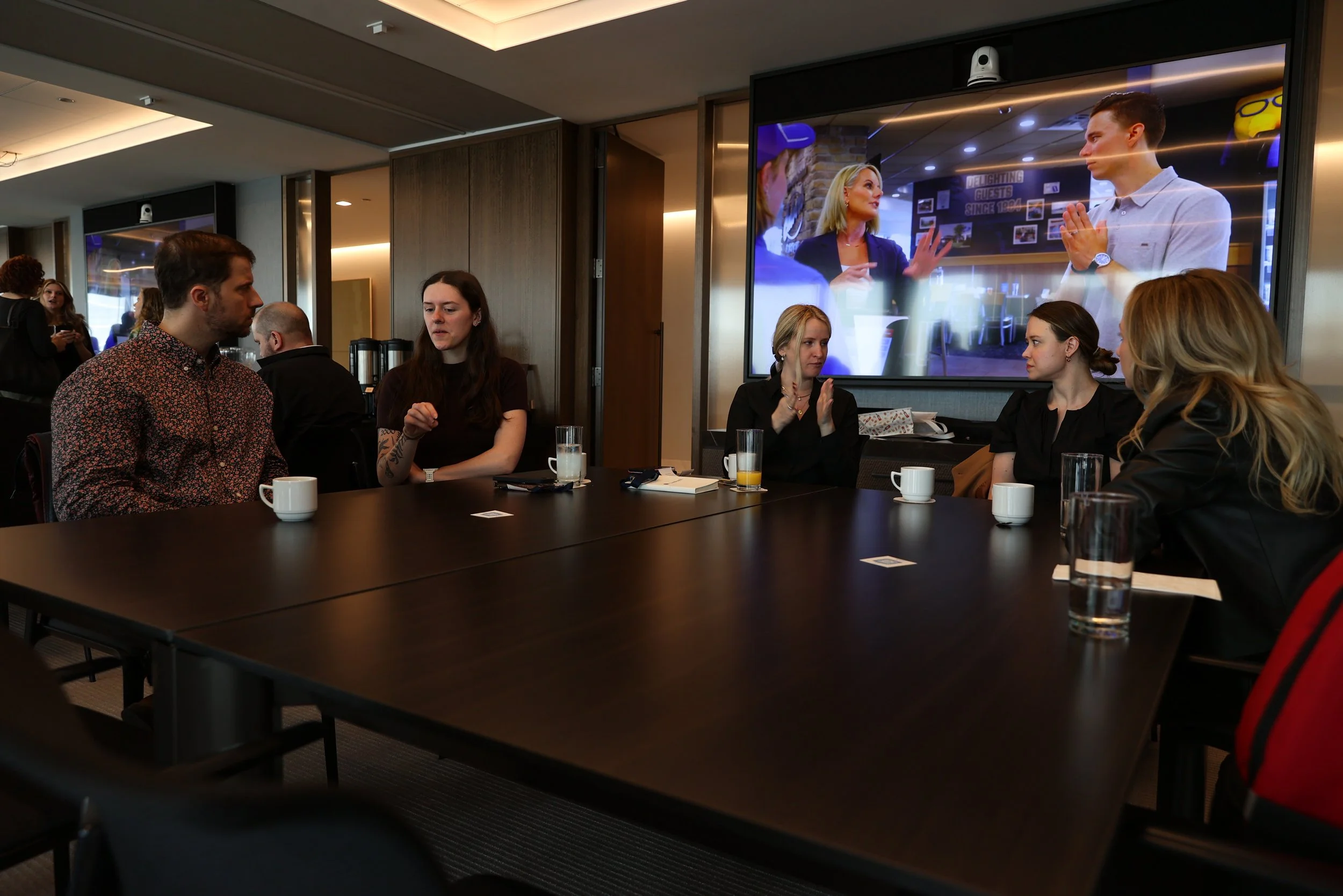 Group of five people sitting at a conference table with drinks, engaged in conversation, in a modern meeting room with large screen displaying a presentation or video conference.