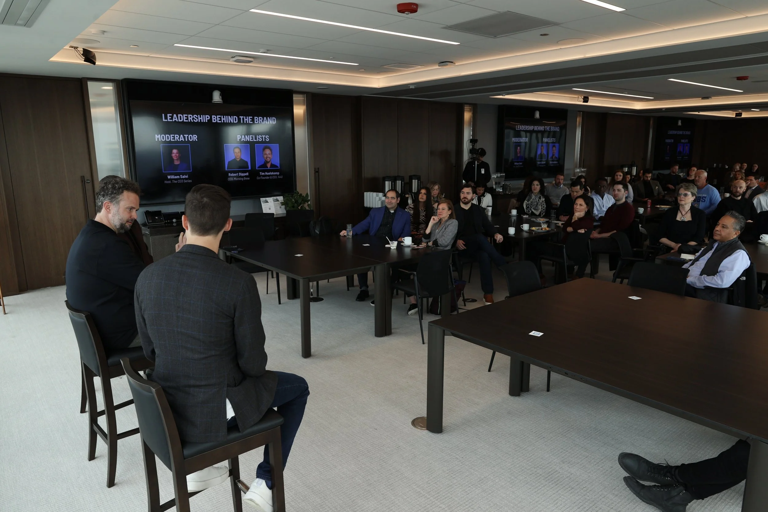 A business conference or panel discussion in a modern conference room with an audience listening to speakers. A large screen behind the speakers displays the event title 'Leadership Behind the Brand', with photos and titles of the moderator and panel