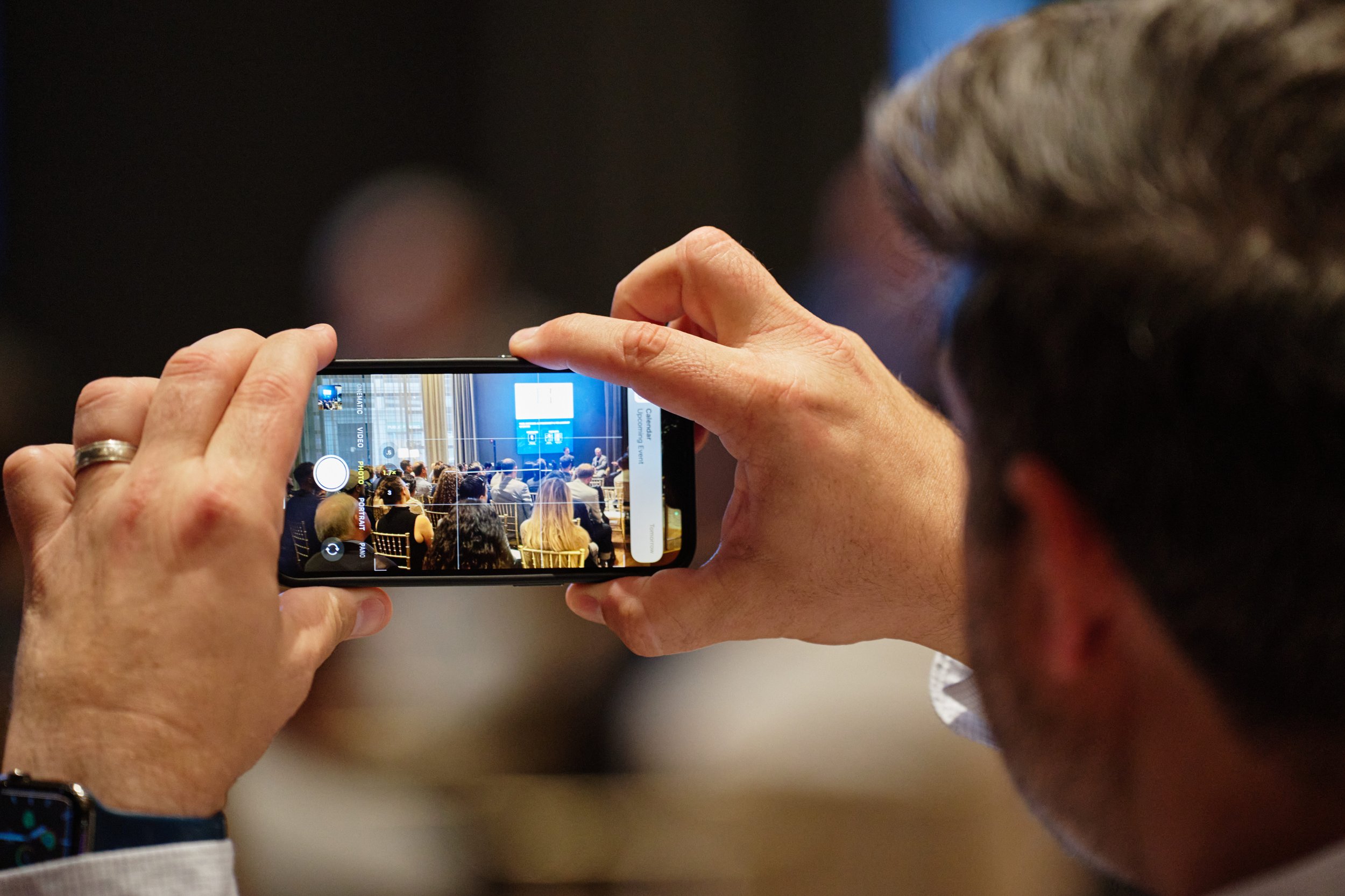 A person taking a photo of a conference or seminar with a smartphone, showing seated audience and a speaker at the front.