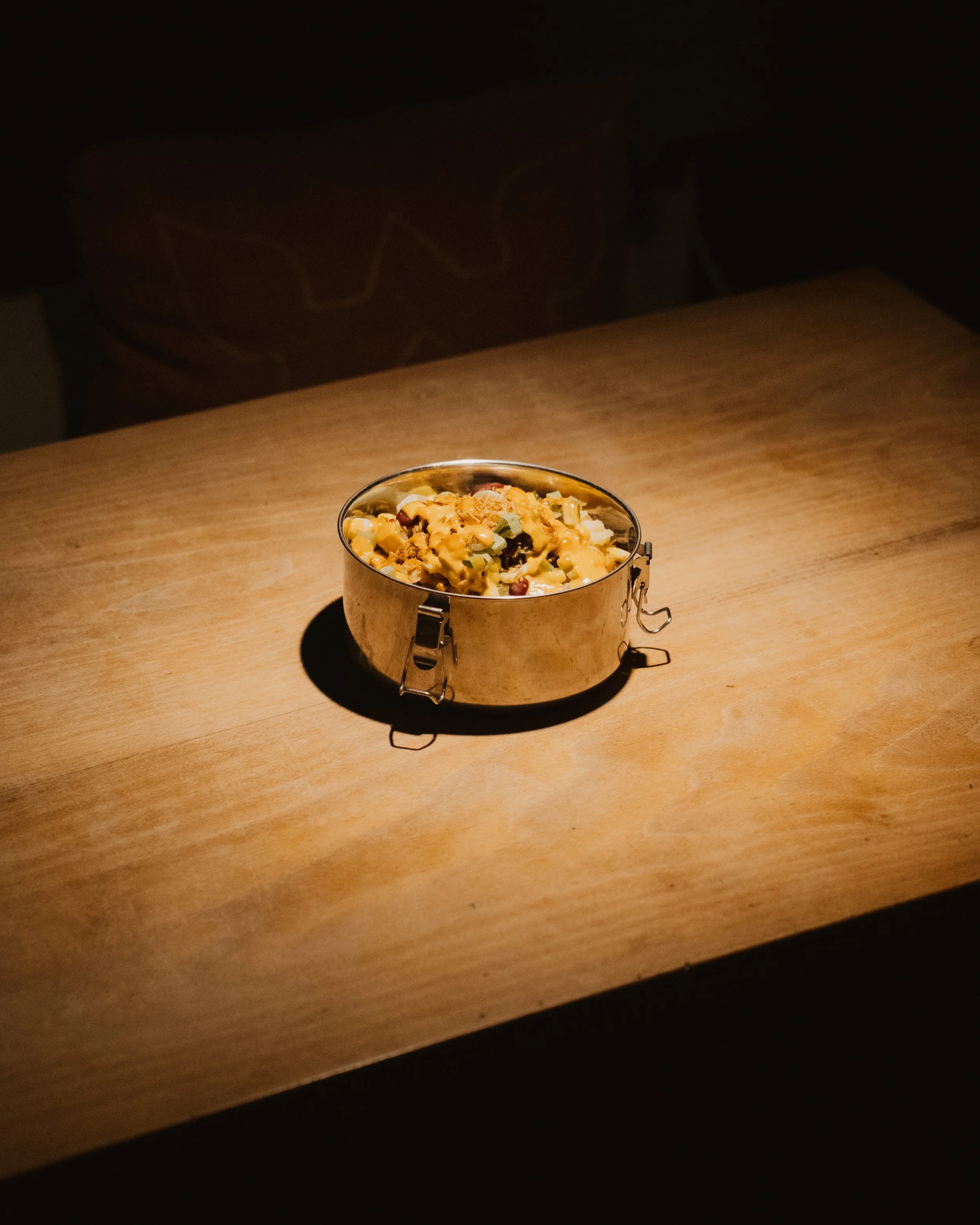 A bowl of pasta salad on a wooden table, illuminated by a spotlight in a dark room.