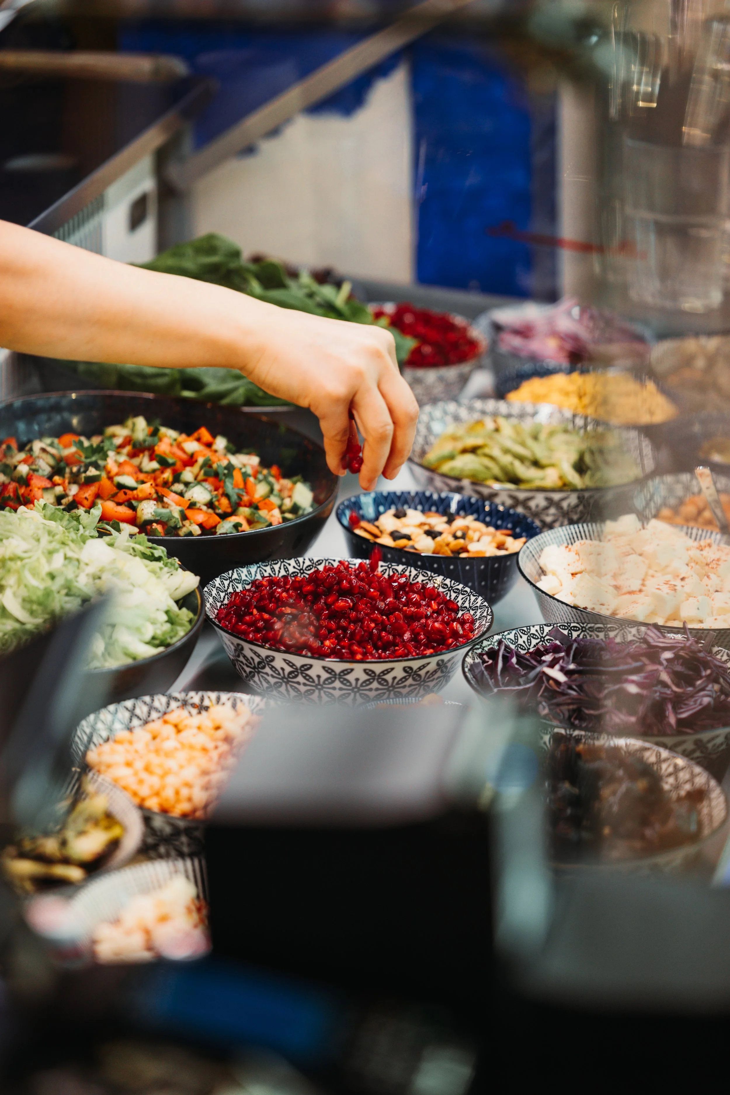 Hand reaching for pomegranate seeds in a bowl at a salad bar with various vegetables and toppings.