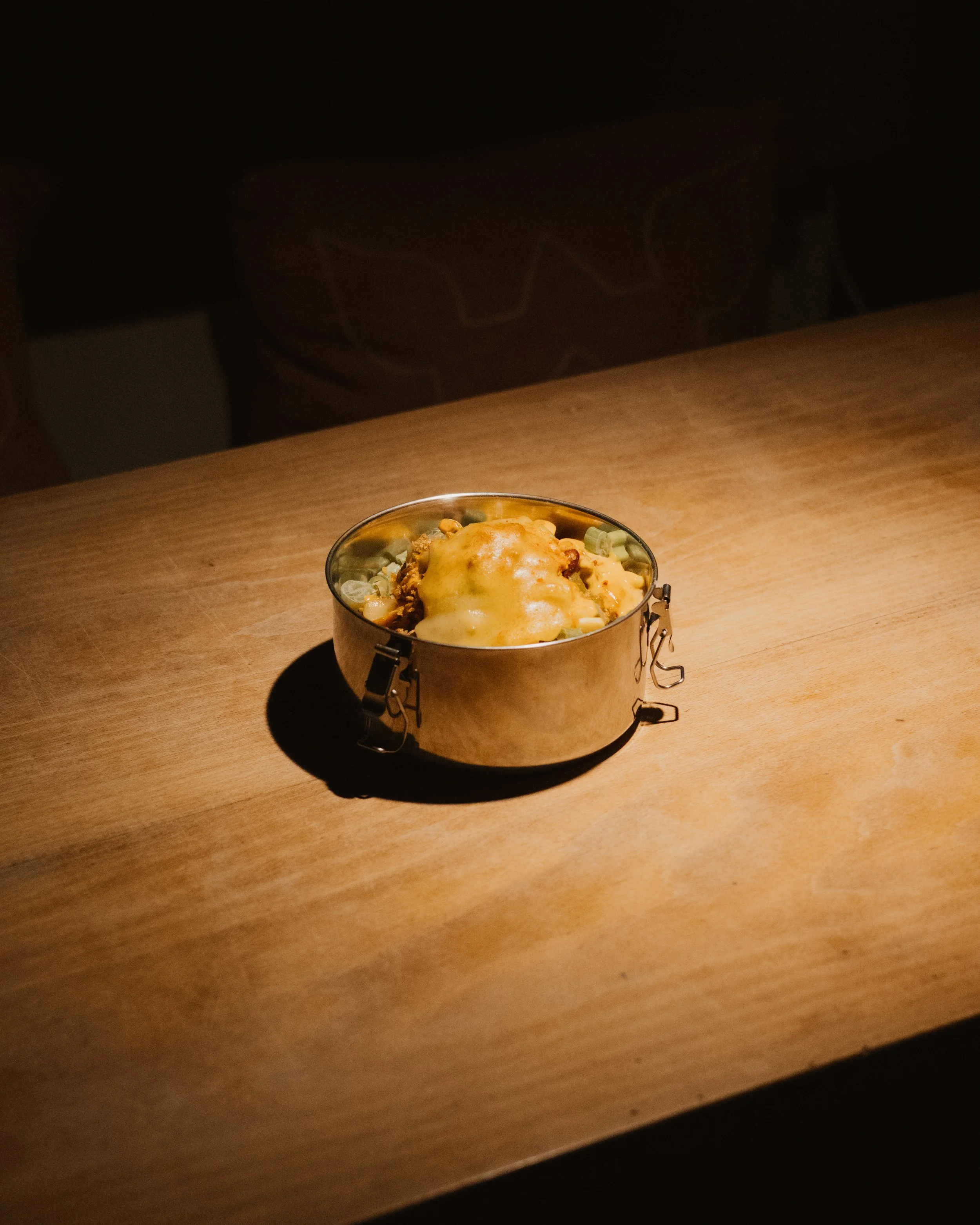 A small round metal container filled with food, topped with melted cheese, placed on a wooden table in a dimly lit setting.