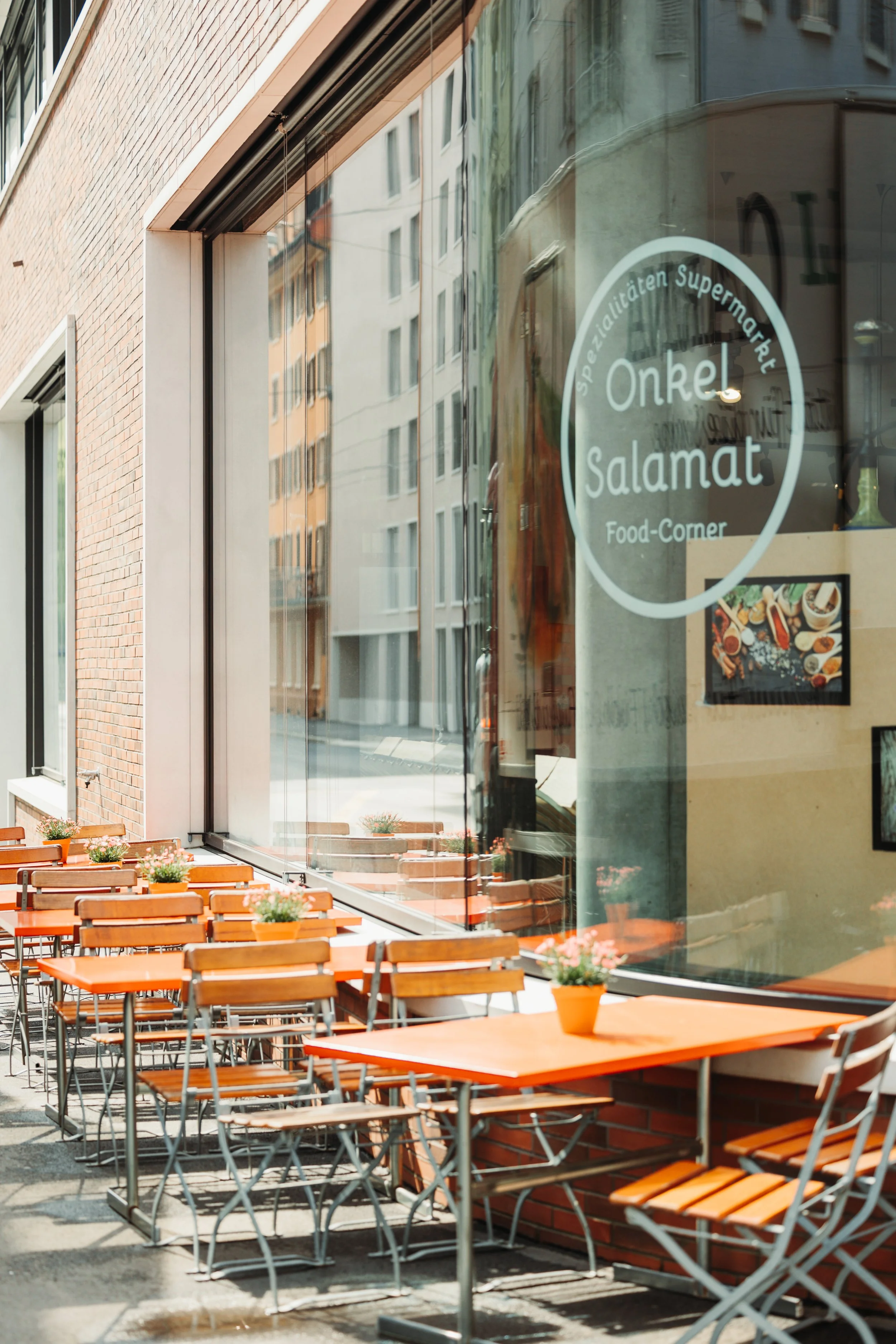 Empty outdoor seating area with orange tables and chairs outside a restaurant named 'Onkel Salamat' with potted plants on the tables and building reflections on the glass window.