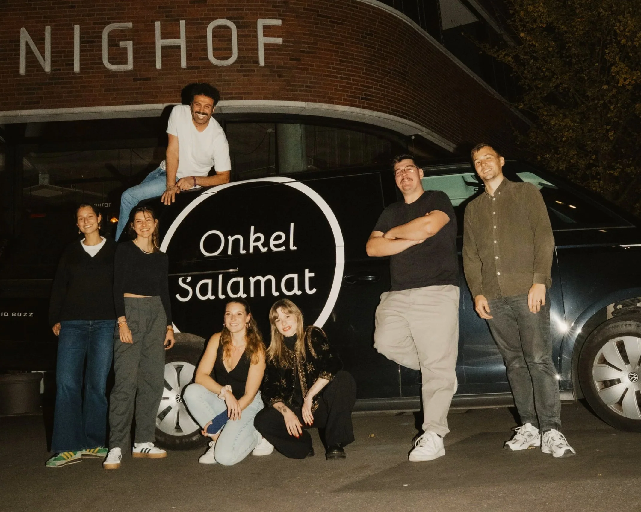 A group of seven young adults posing in front of a black van with white text that reads "Onkel Salamat." The group is standing and sitting on the pavement at night, with a brick building and trees in the background.