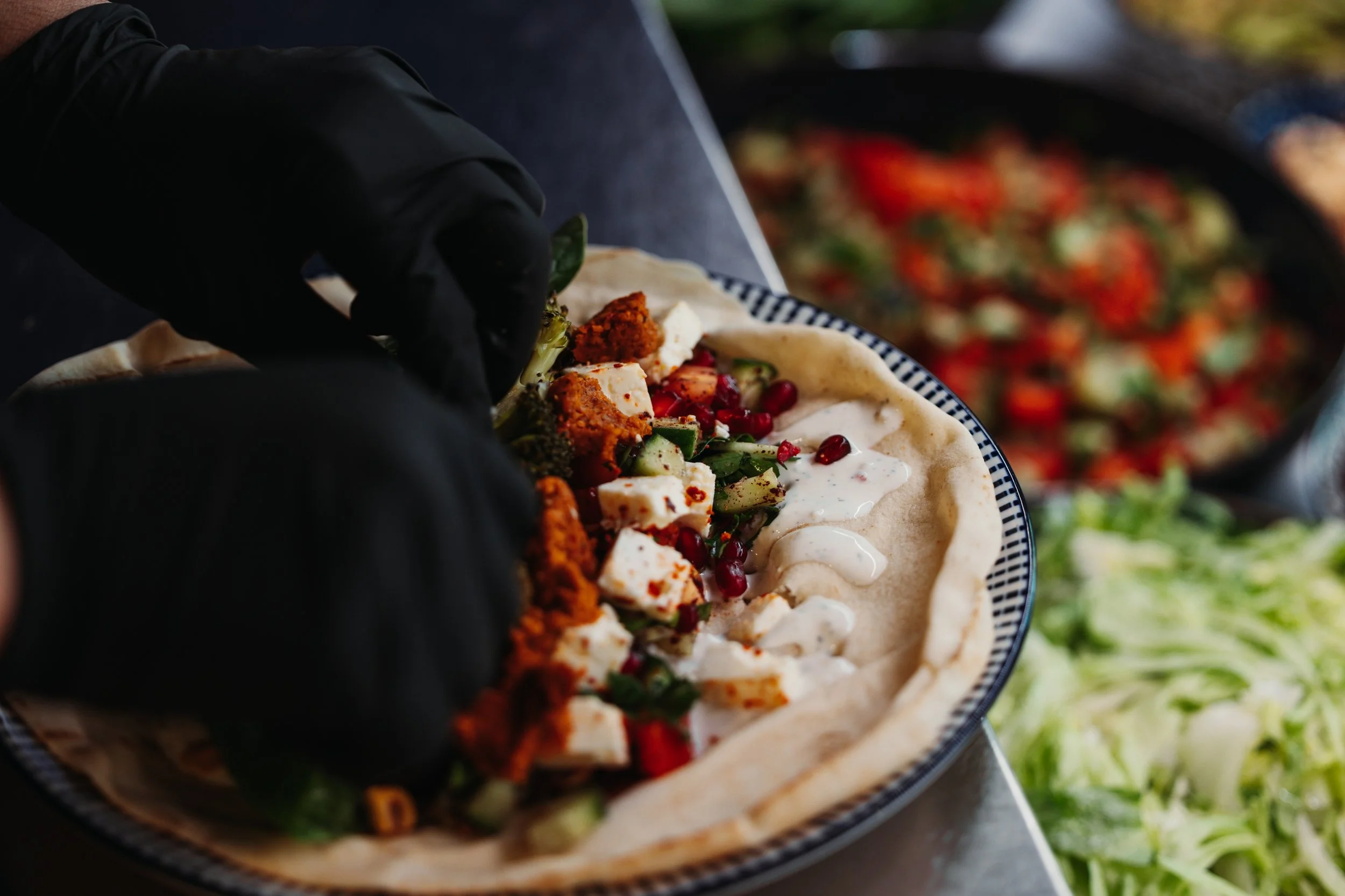 Person wearing black gloves preparing a Greek salad in a bowl, with vegetables, feta cheese, and dressing. Various bowls of salad ingredients in the background.