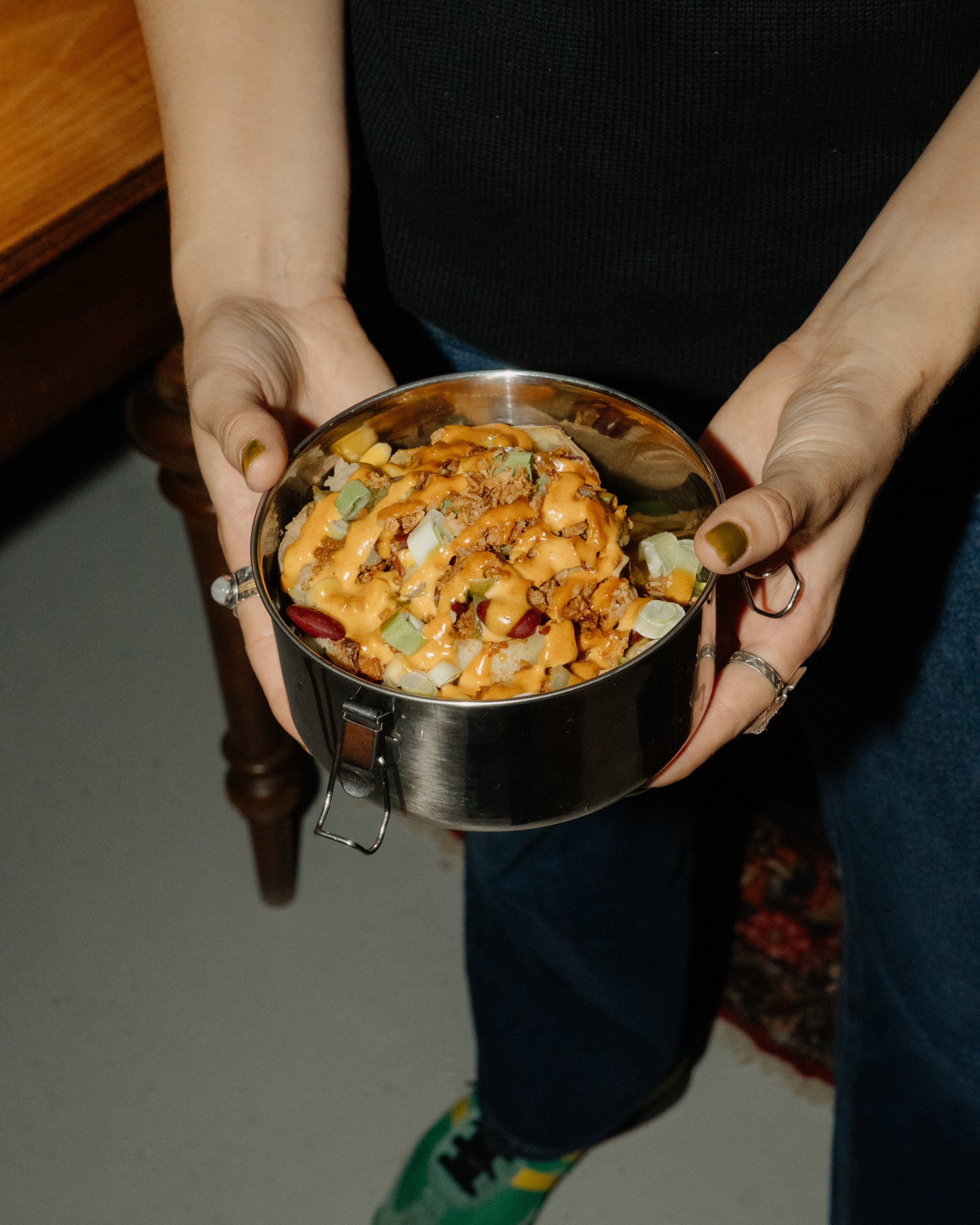 Person holding a metal bowl filled with food, topped with sauce and garnished with chopped green onions, with a wooden piece and a colorful carpet visible on the floor.