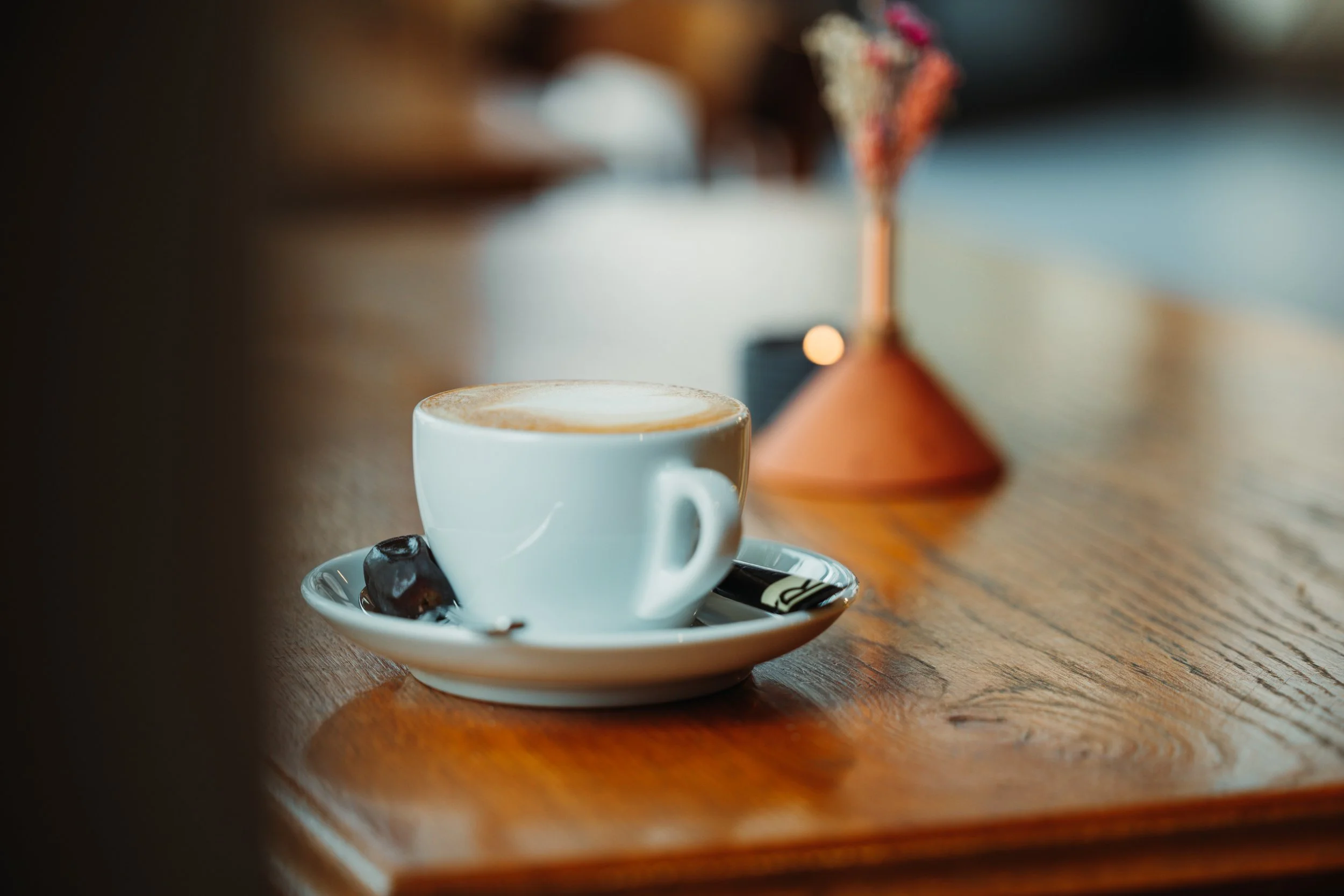 A white cup of coffee or latte on a saucer with a piece of chocolate and a spoon, placed on a wooden table.