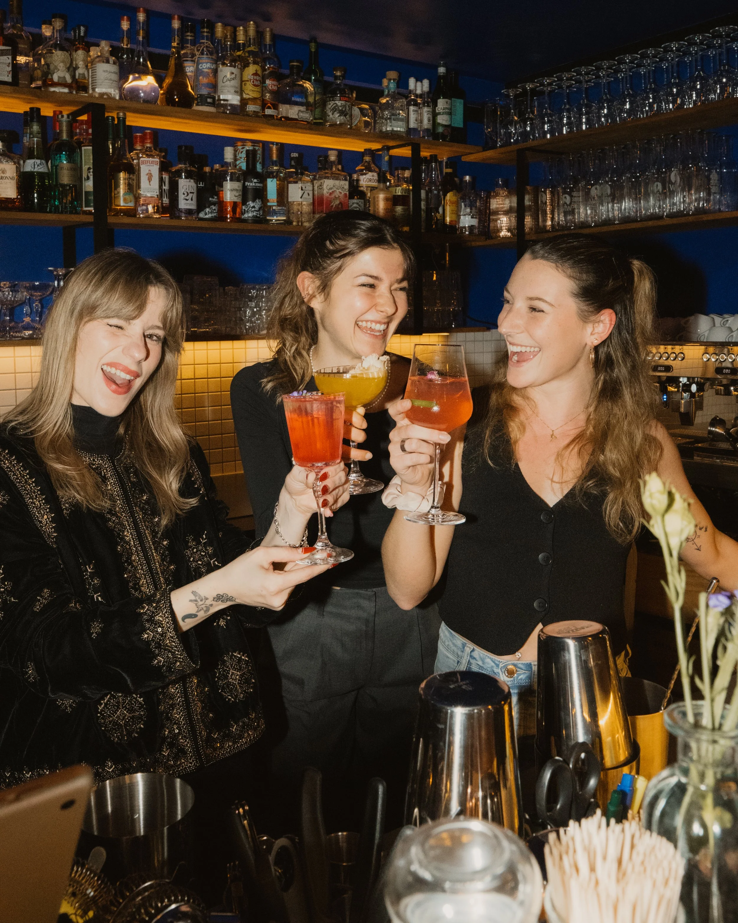 Three women enjoying drinks and celebrating at a bar, smiling and holding colorful cocktails.