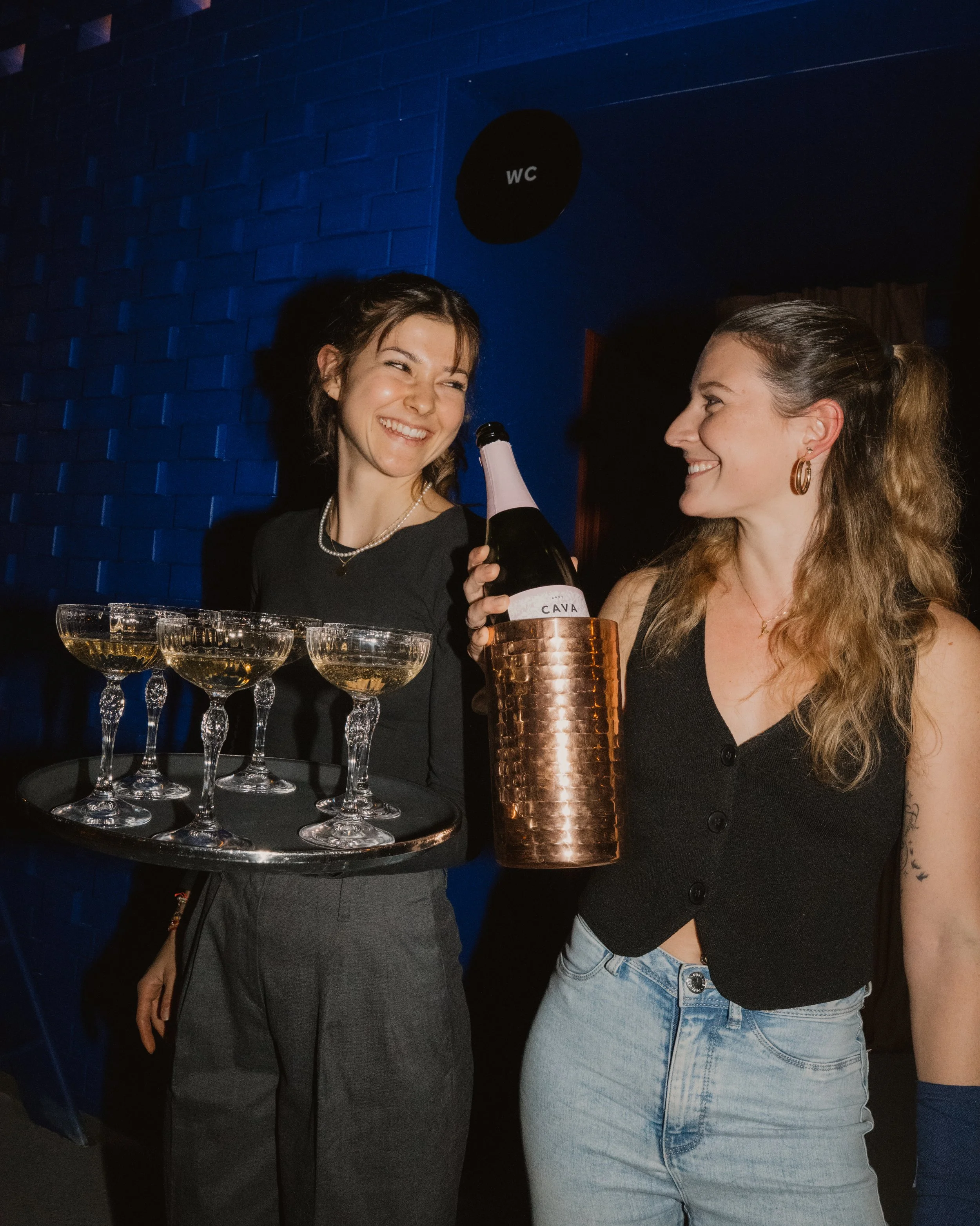 Two women smiling and holding glasses of champagne, one with a tray of four glasses, in a dimly lit party setting.