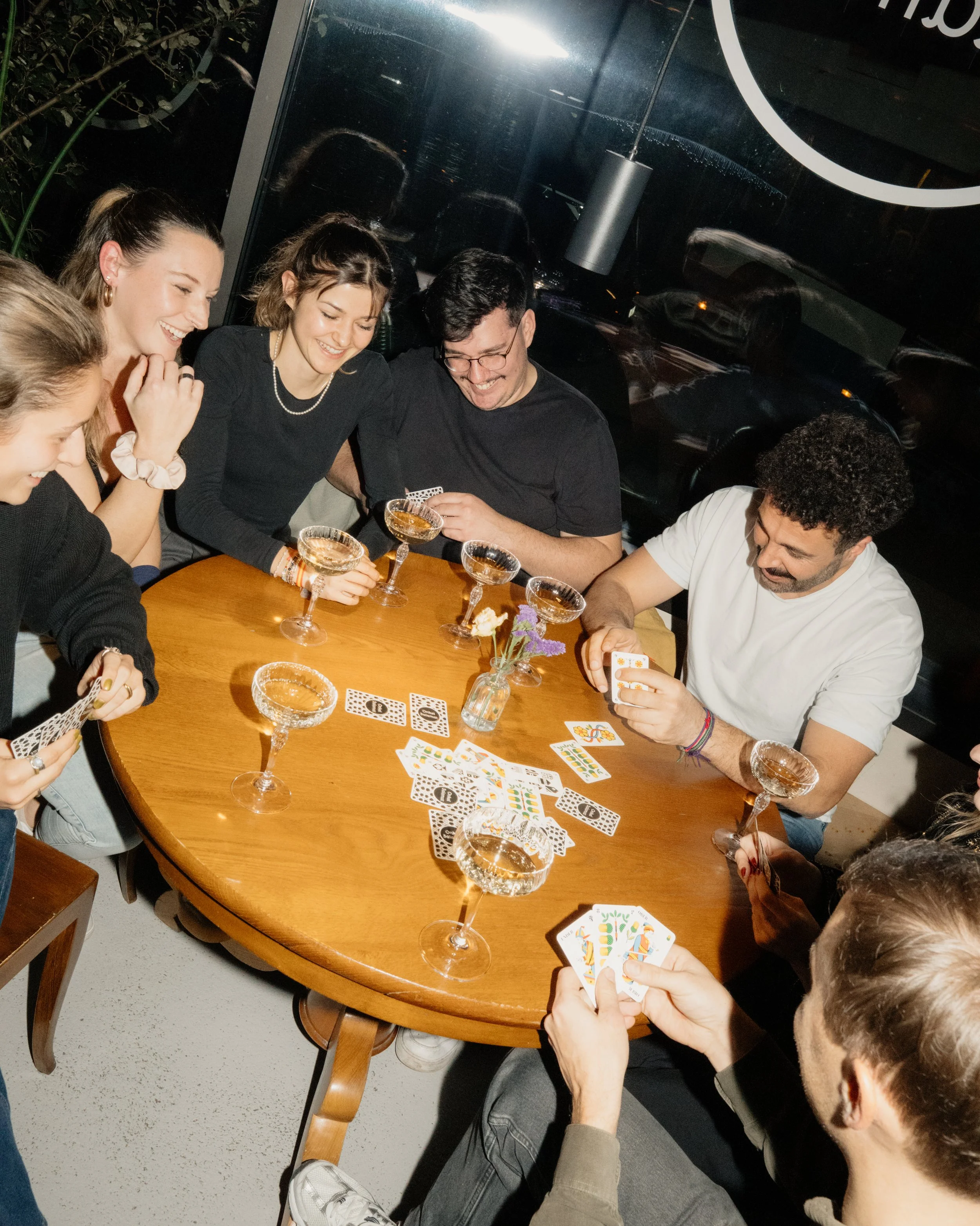 Seven people gathered around a wooden table playing cards and laughing. There are six champagne glasses on the table, and a small vase with purple and white flowers.