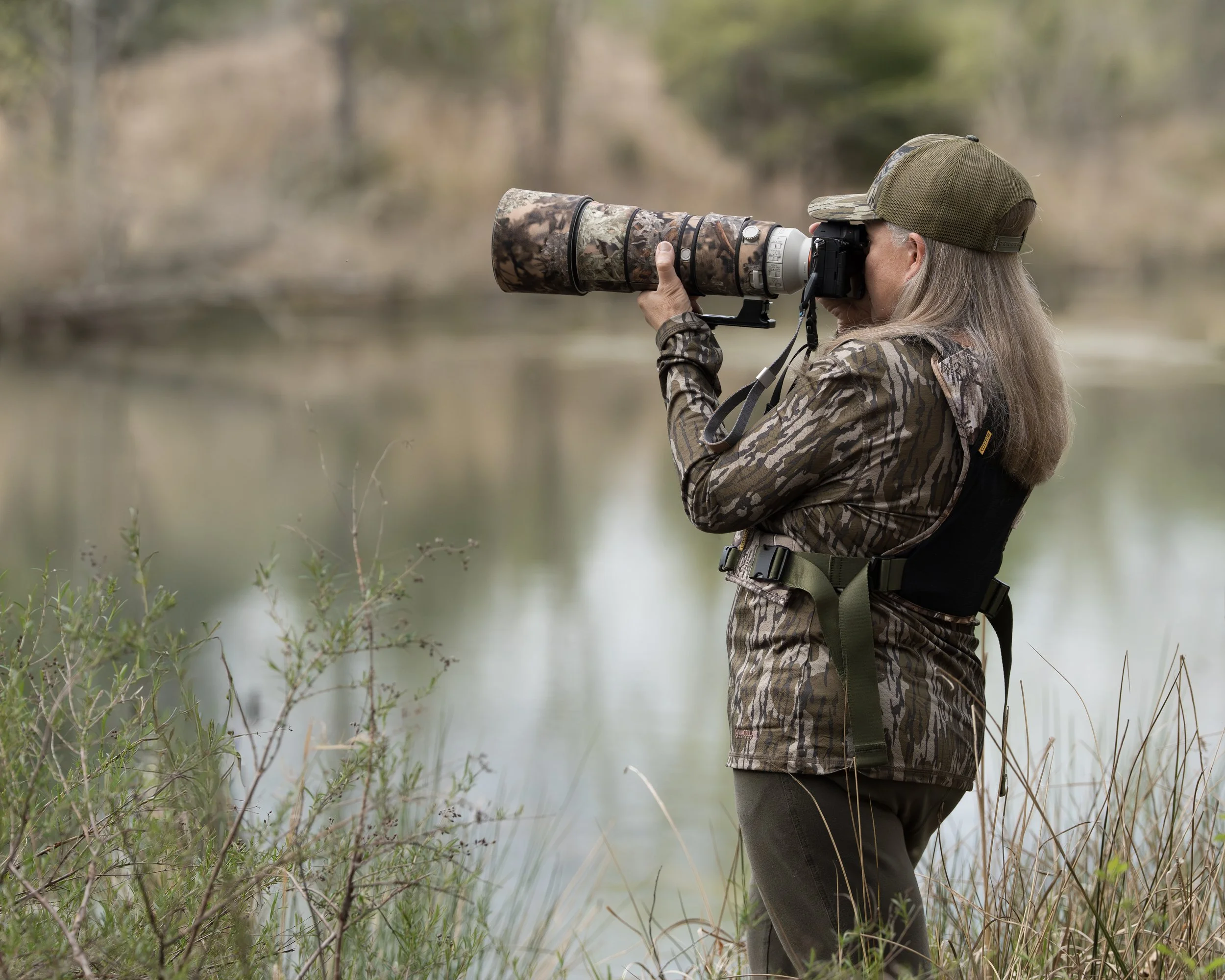 A woman in camouflage clothing and a cap, using a telephoto lens with a camera, standing by a lake in a wooded area.