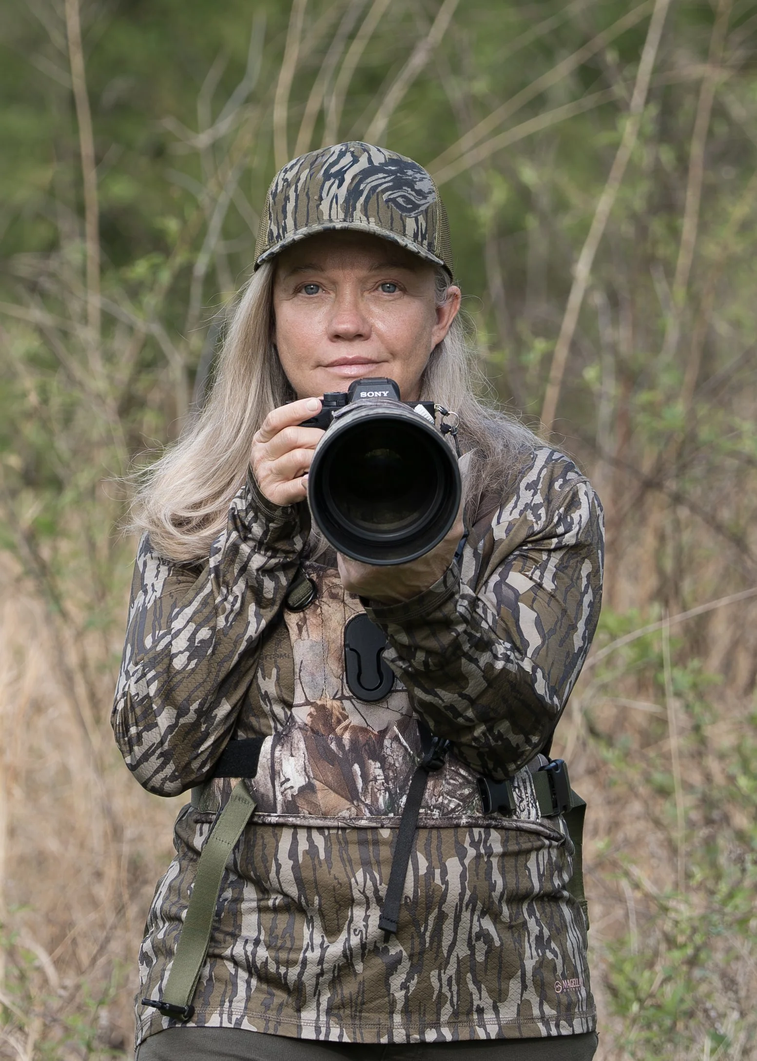 A woman in camouflage clothing and a camo cap taking a photo with a Sony camera in a forested area.