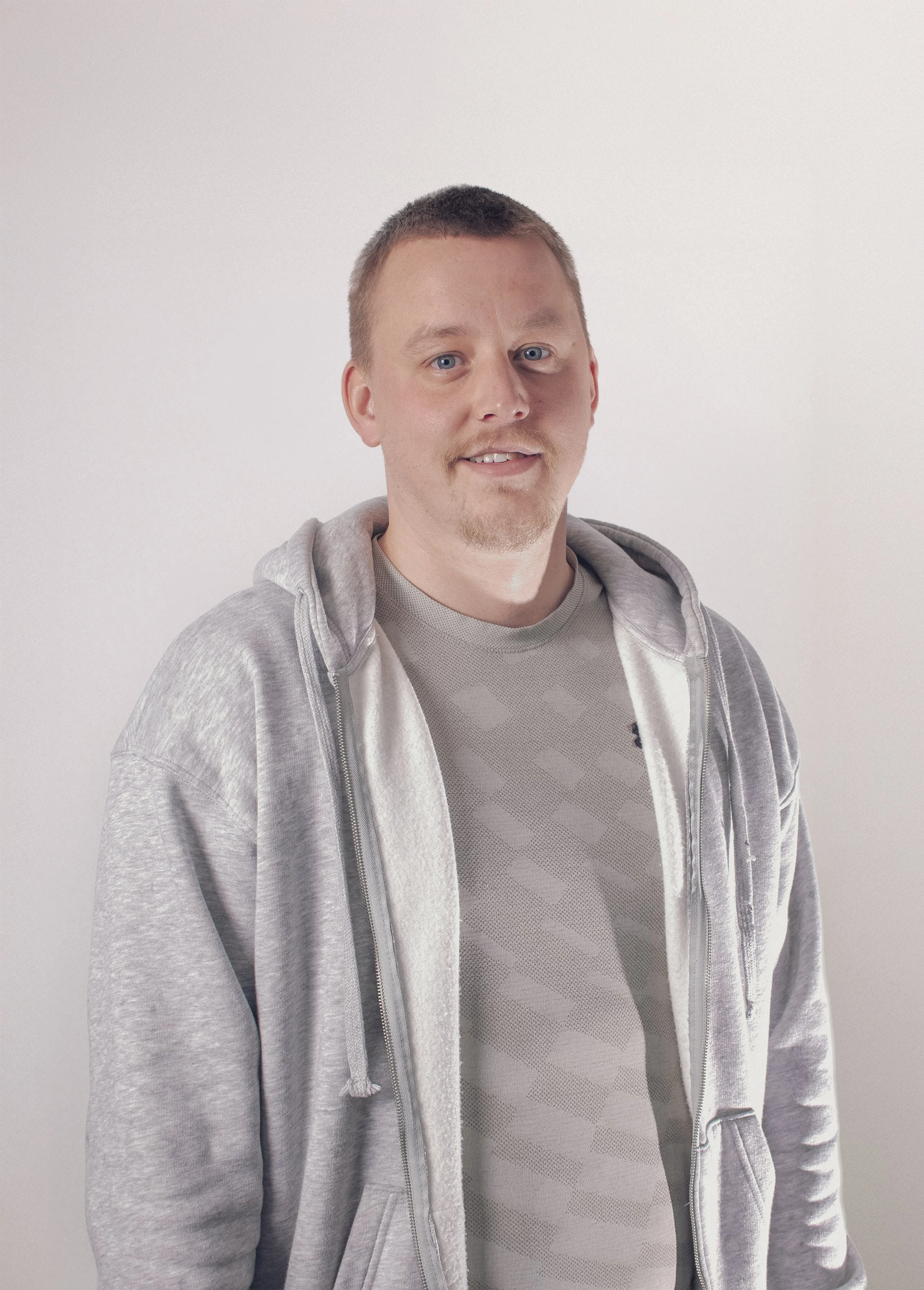 A man with short hair, blue eyes, and a light beard smiling, wearing a grey zip-up hoodie over a grey shirt, standing against a plain white background.
