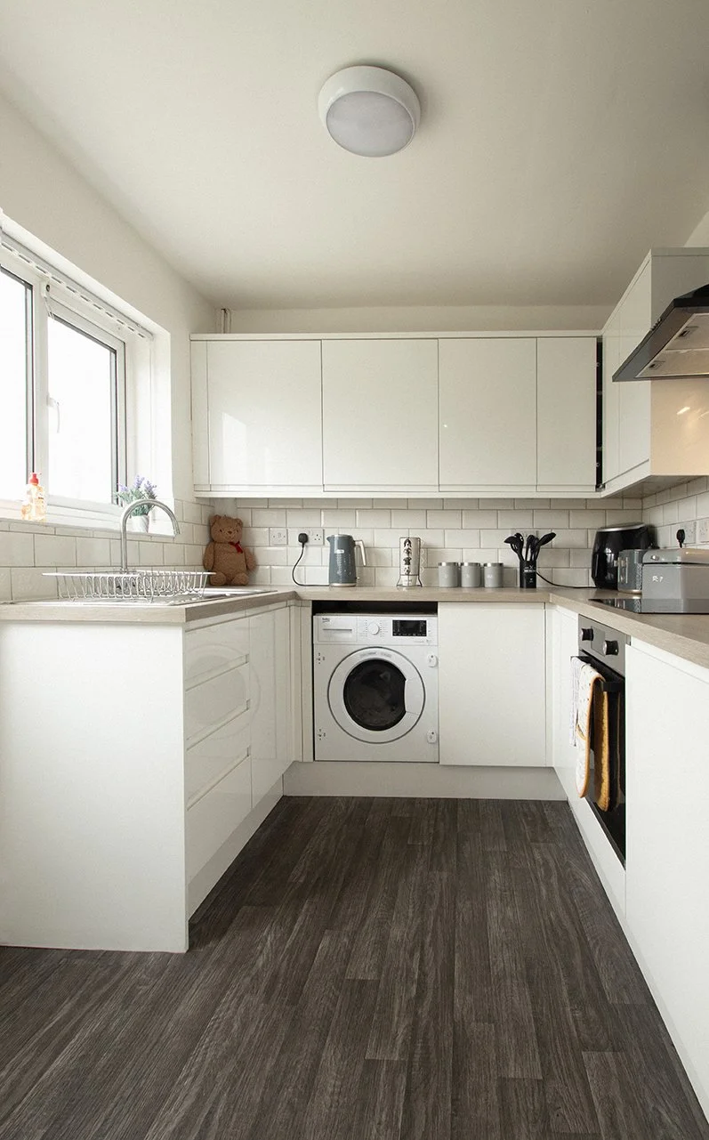 A modern kitchen with white cabinets, a window, a round ceiling light, and dark wooden flooring. It features a washing machine, a sink, and various small appliances on the countertops.