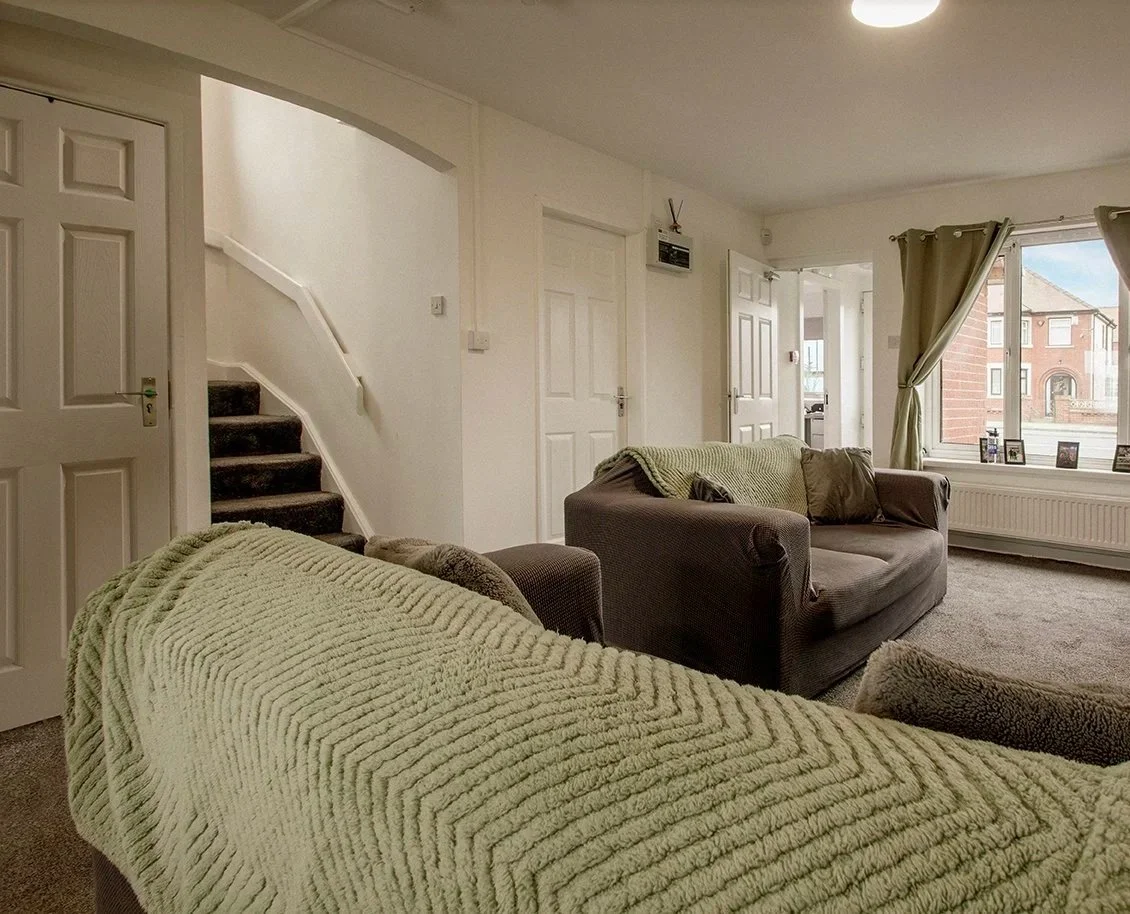 Living room with two sofas, one with a green blanket, near a large window with curtains and photos on the windowsill. A staircase to the left and a view into an adjacent room are visible.