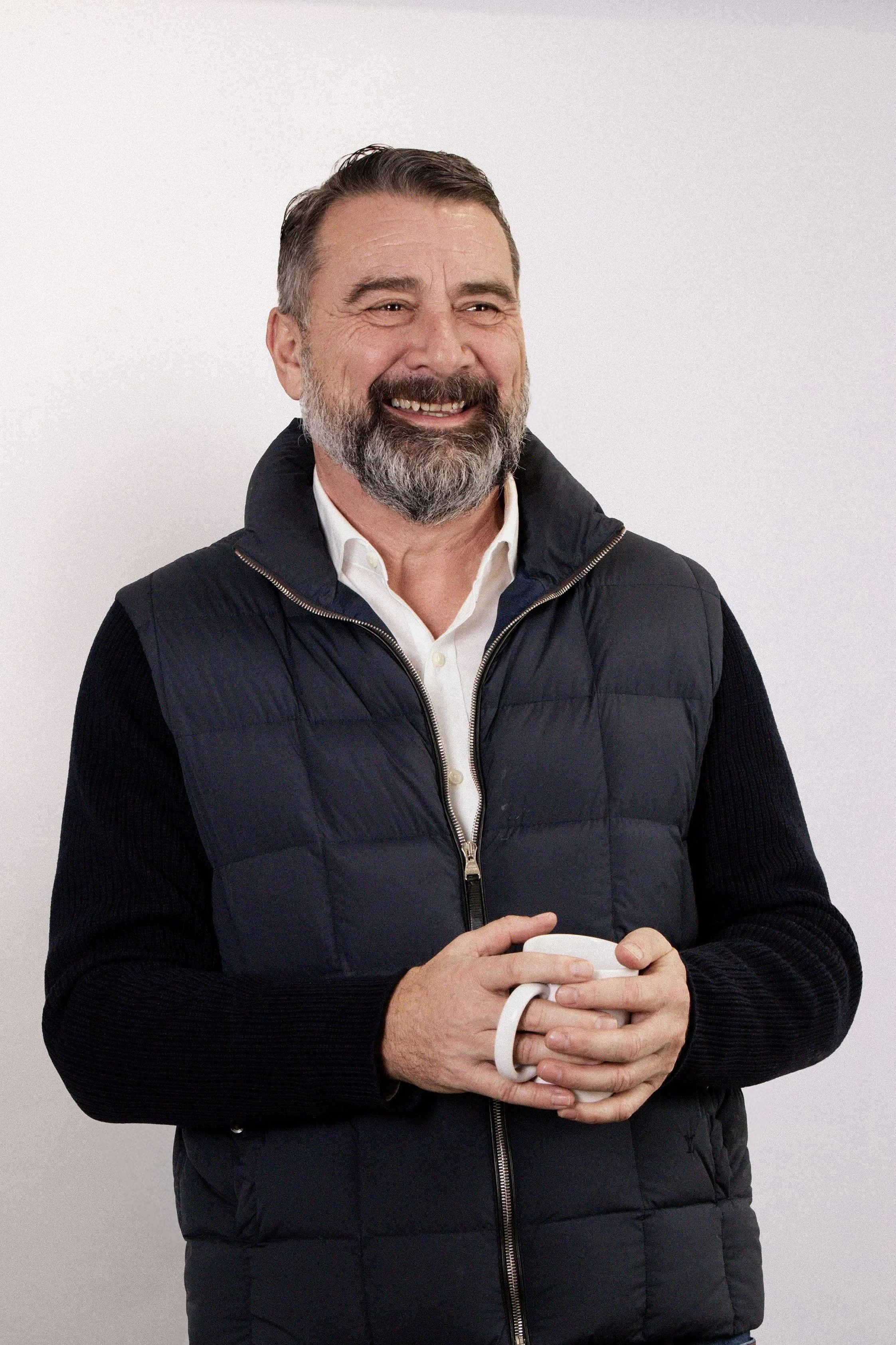 A smiling middle-aged man with a beard, wearing a white shirt and a black quilted vest, holding a white mug, standing against a plain light background.