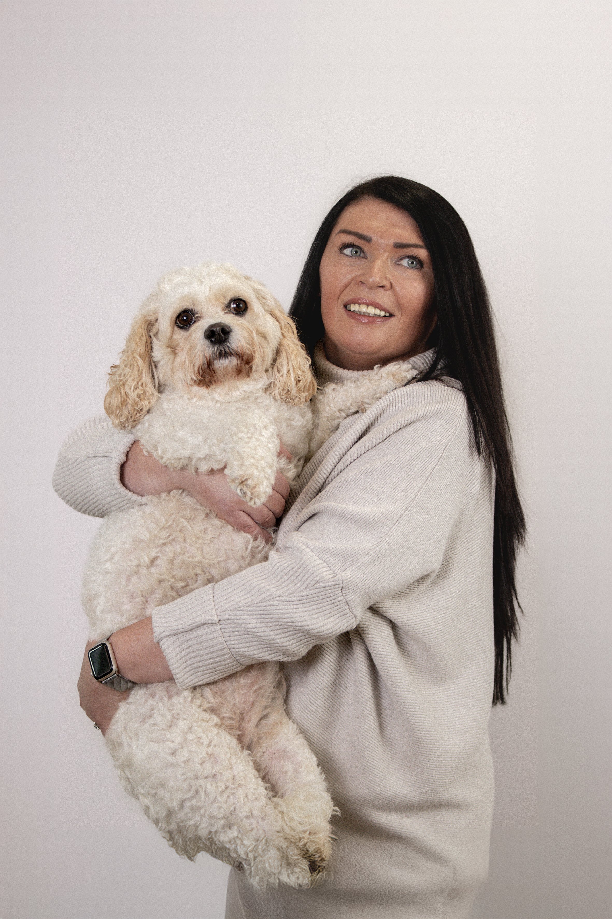 Woman with long dark hair, wearing a beige sweater, holding a fluffy white dog with curly fur against a plain white background.